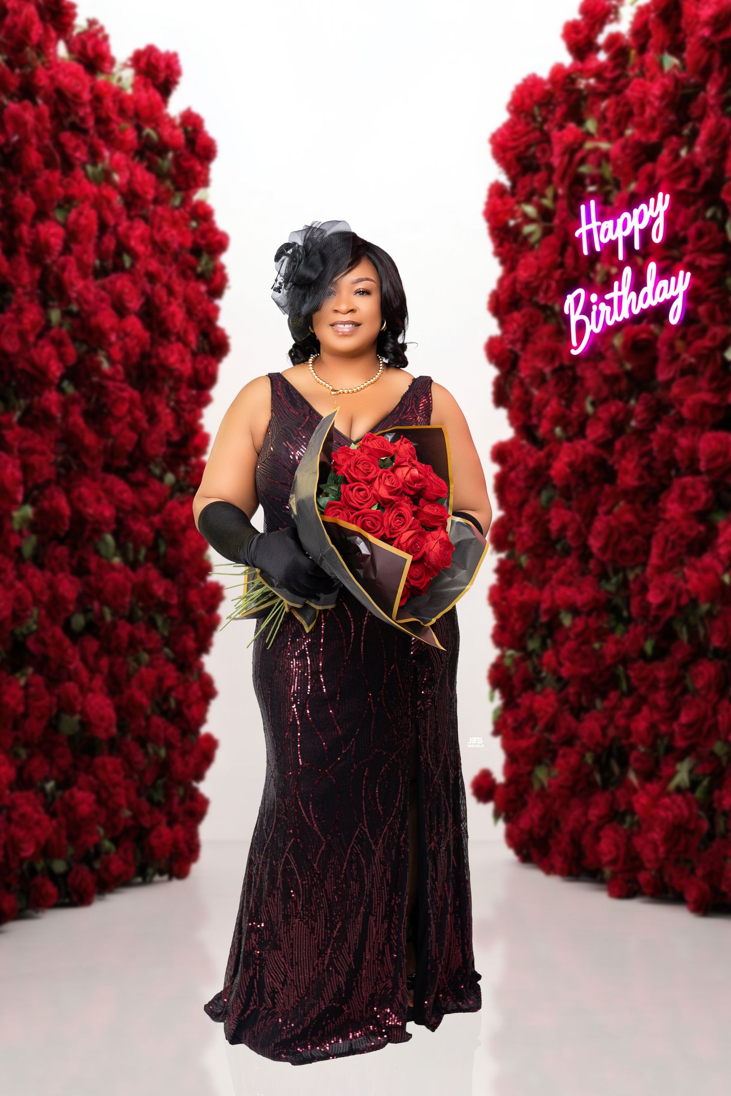 Woman in burgundy sequin gown holds red roses between rose walls with a Happy Birthday neon sign.