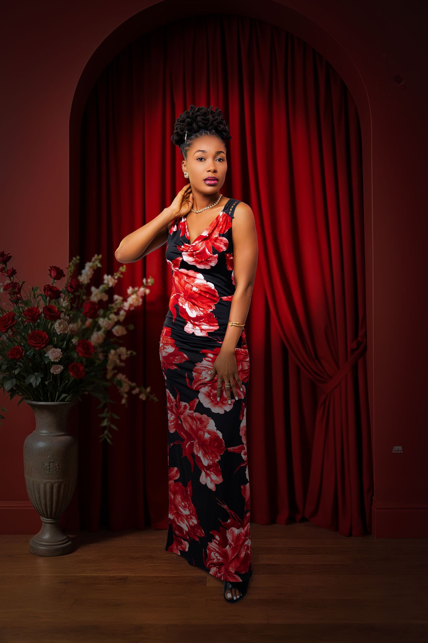 Beautiful woman in black and red floral maxi dress poses before red curtain arch with roses.