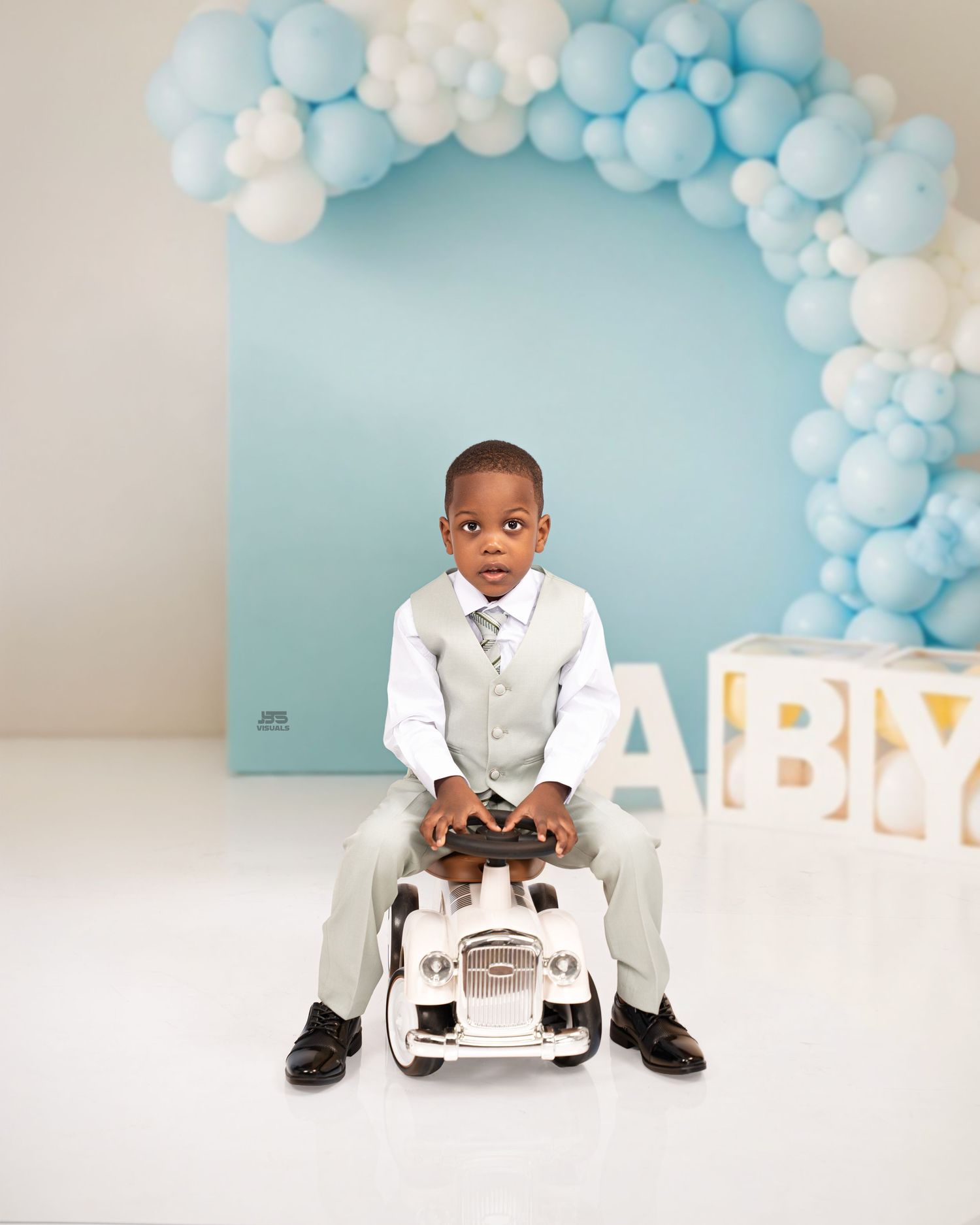 Young boy in gray suit sits on toy car with blue balloon arch backdrop and BABY letters.