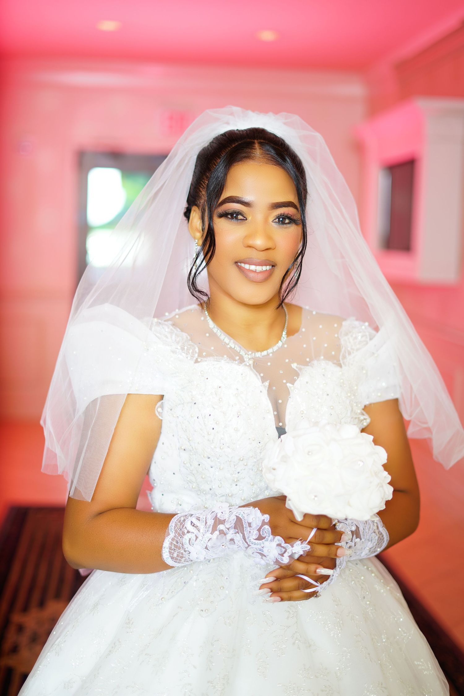 Beautiful bride in white lace ball gown with veil and gloves posing in a pink-lit room.