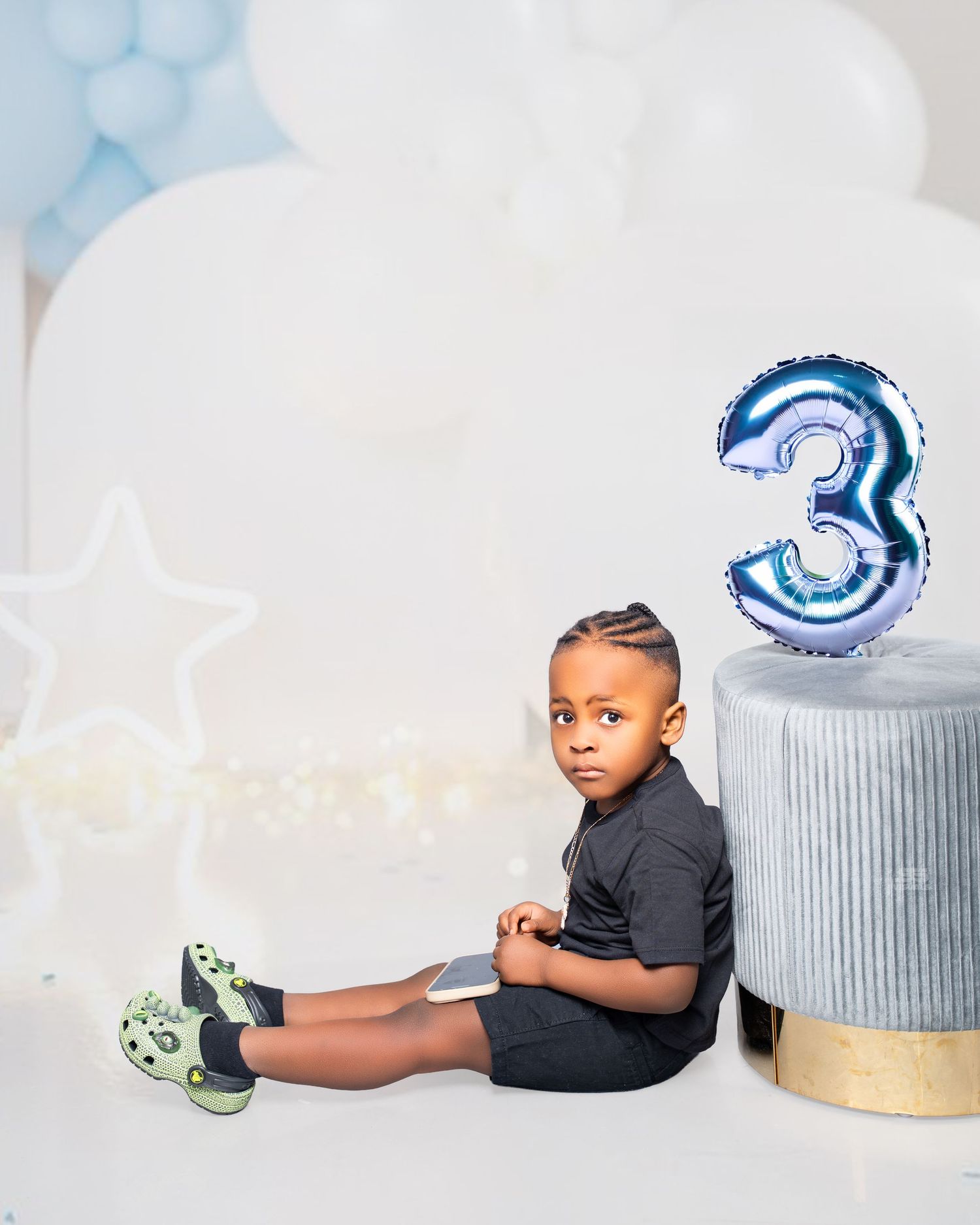 Young Black boy sitting beside a number 3 balloon in blue birthday party studio setup.