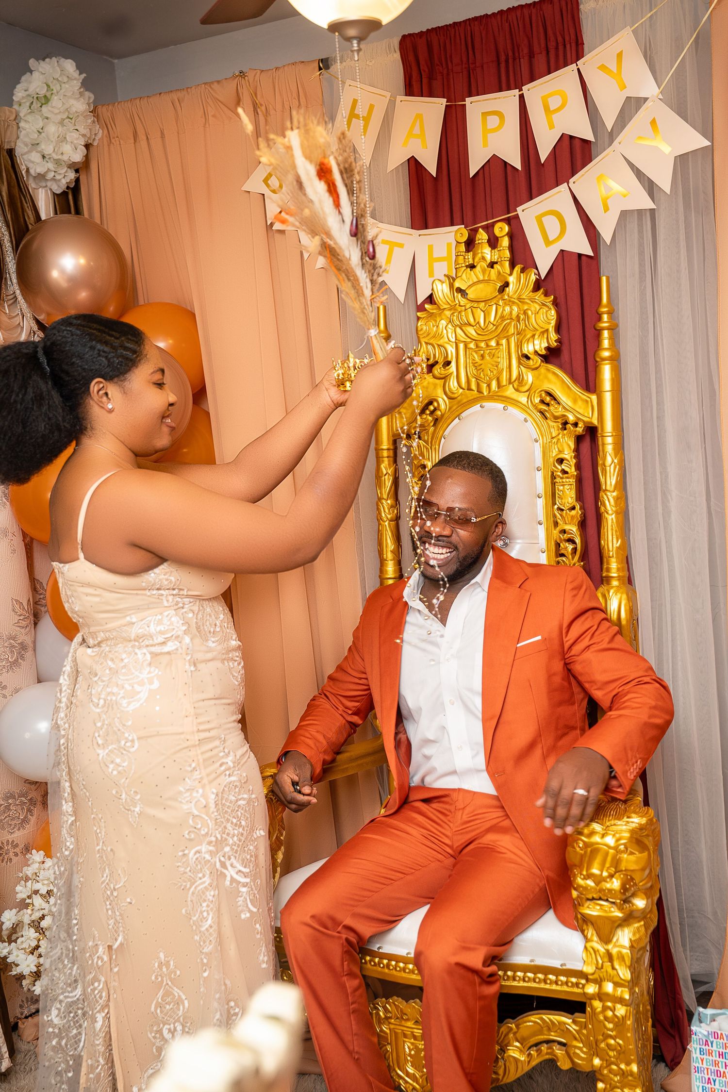 Man in orange suit sits on gold throne as woman celebrates his birthday party.