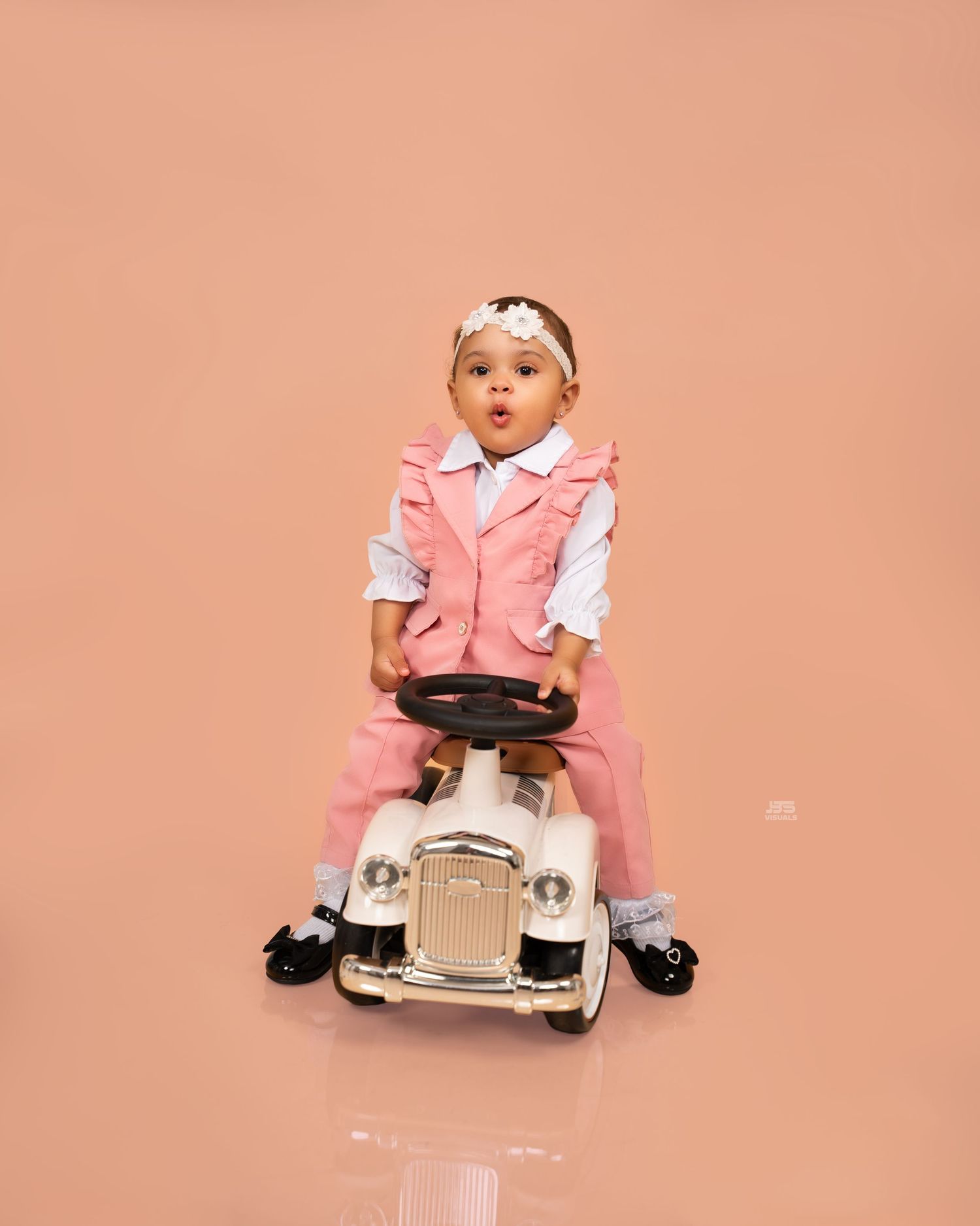 Adorable baby girl in pink suit riding a vintage toy pedal car against a soft pink background.