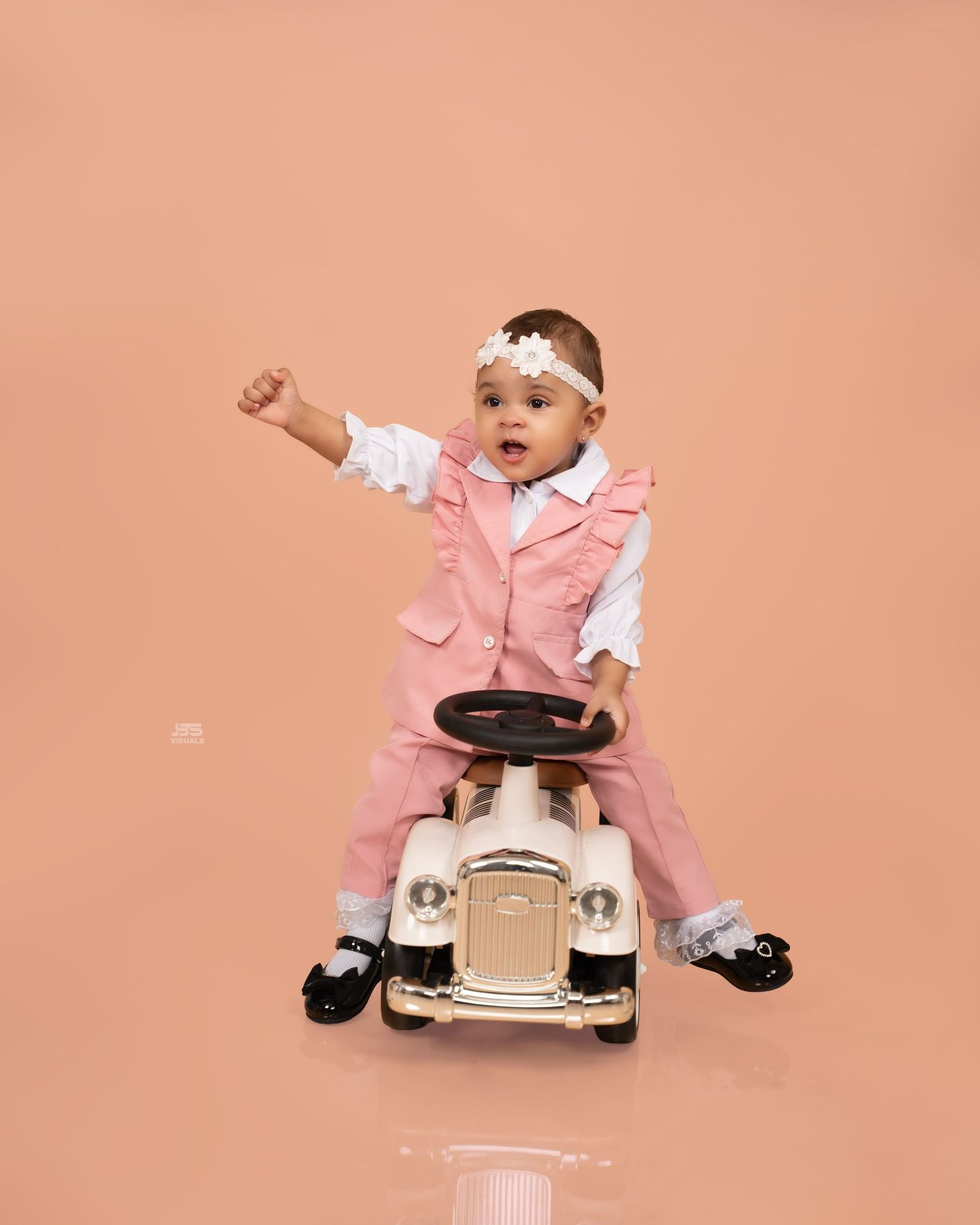 Baby girl in pink suit sits on vintage toy car, fist raised, wearing white floral headband on peach background.