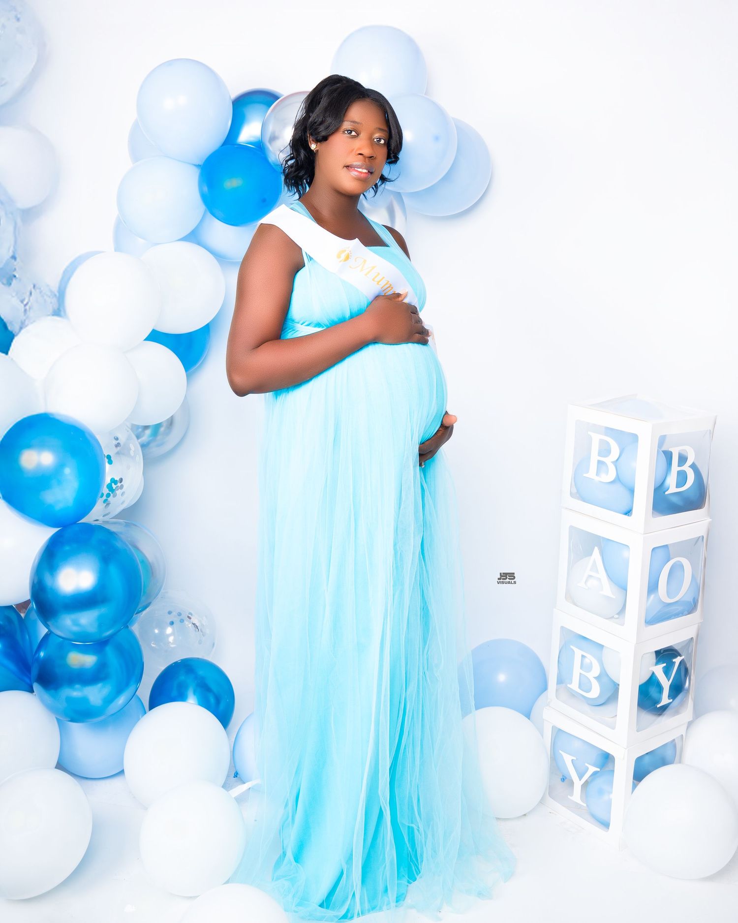 Pregnant woman in light blue tulle gown poses with blue and white balloons and BABY letter boxes.