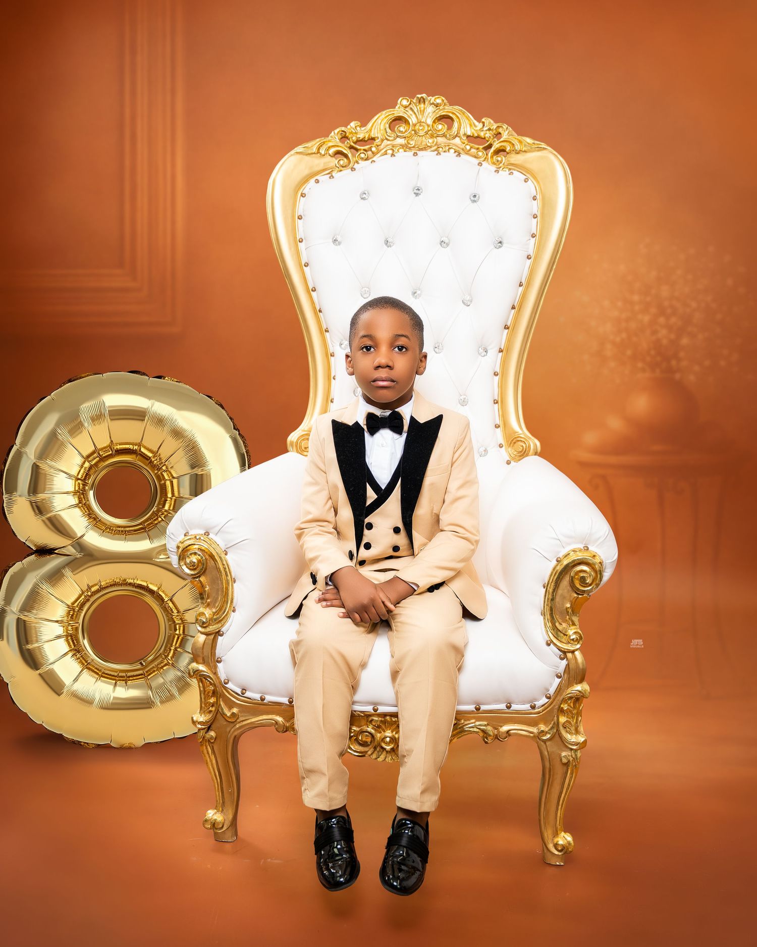 Boy in beige tuxedo sitting on gold throne chair beside number 8 balloon for birthday photos.
