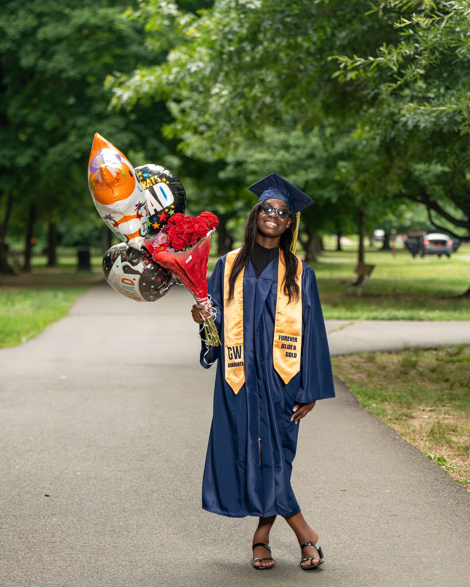 Smiling graduate in navy blue gown holding balloons and flowers in park.