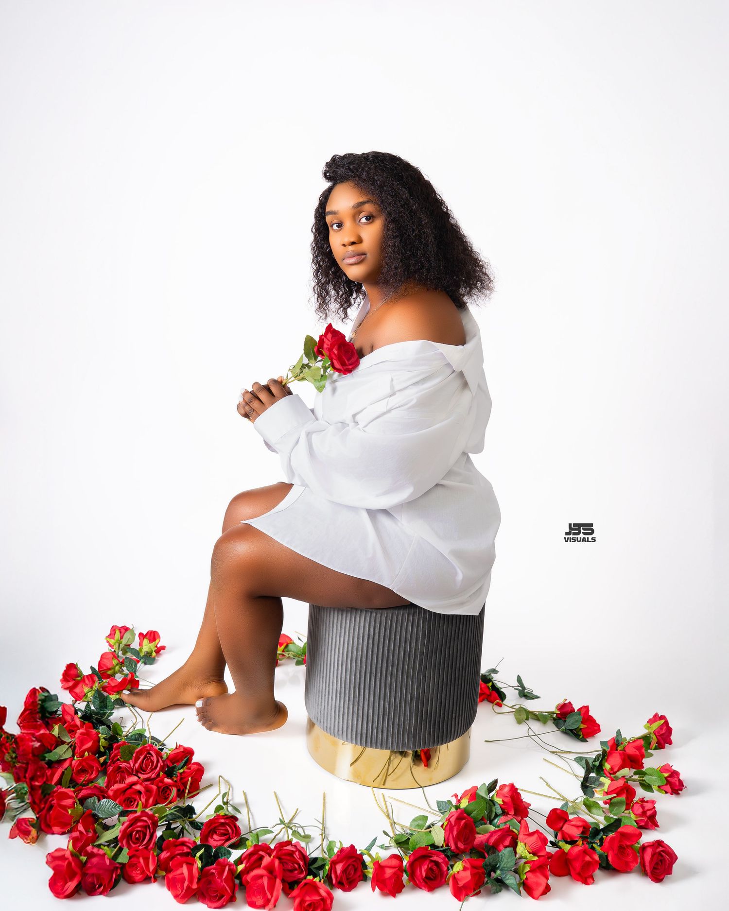 Woman in white off-shoulder top seated on stool surrounded by red roses on white background.