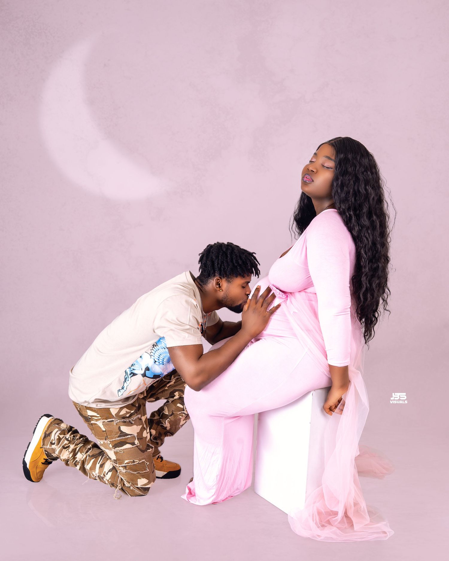 Man kneels kissing pregnant woman's baby bump in pink maternity photo shoot with moon backdrop.