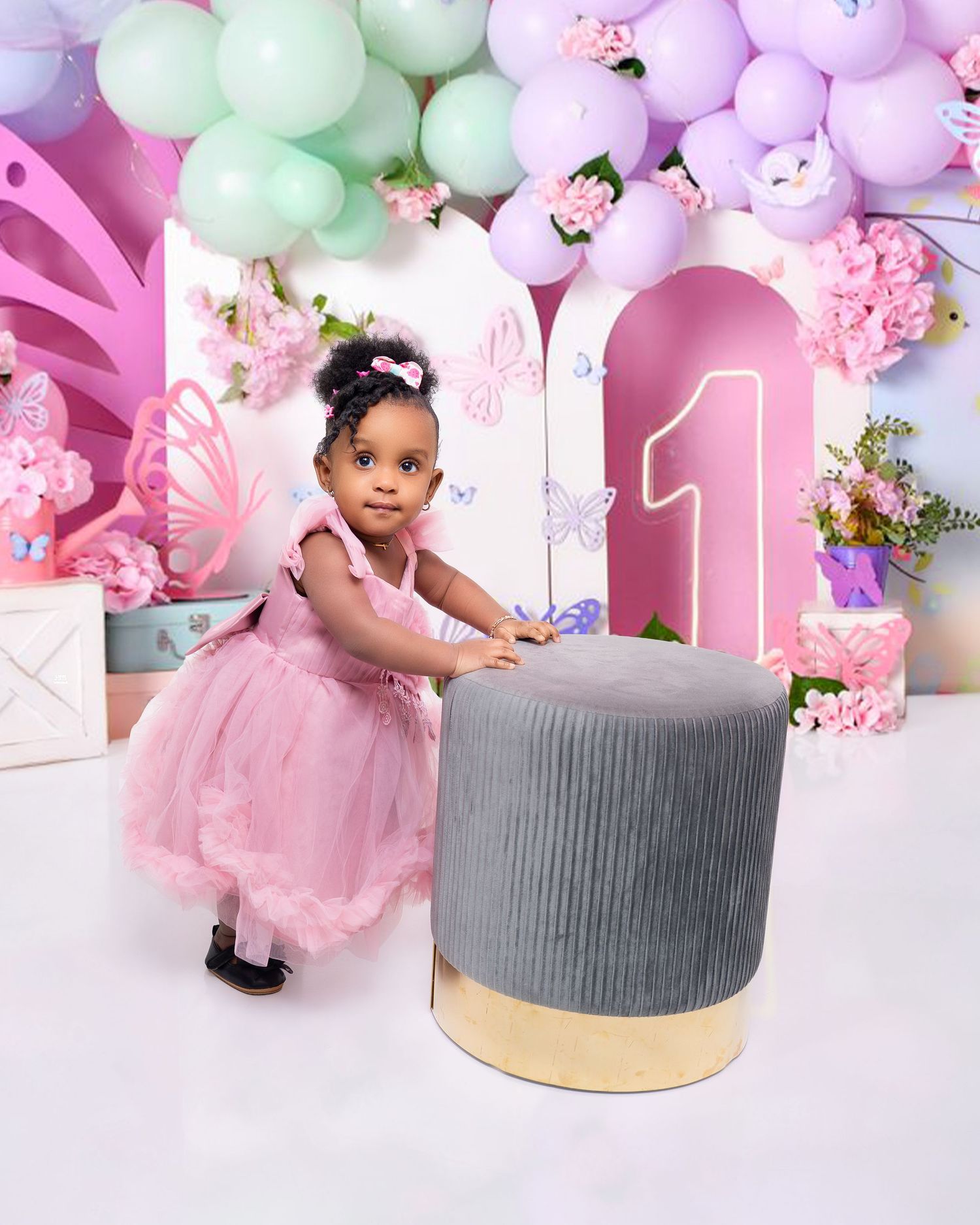 Adorable baby girl in pink tutu dress standing by gray ottoman at butterfly first birthday setup.