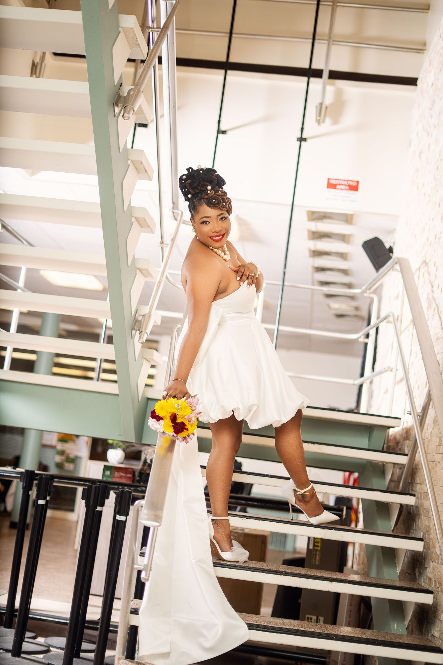 Elegant bride in short white wedding dress holding colorful bouquet while posing on modern interior staircase.