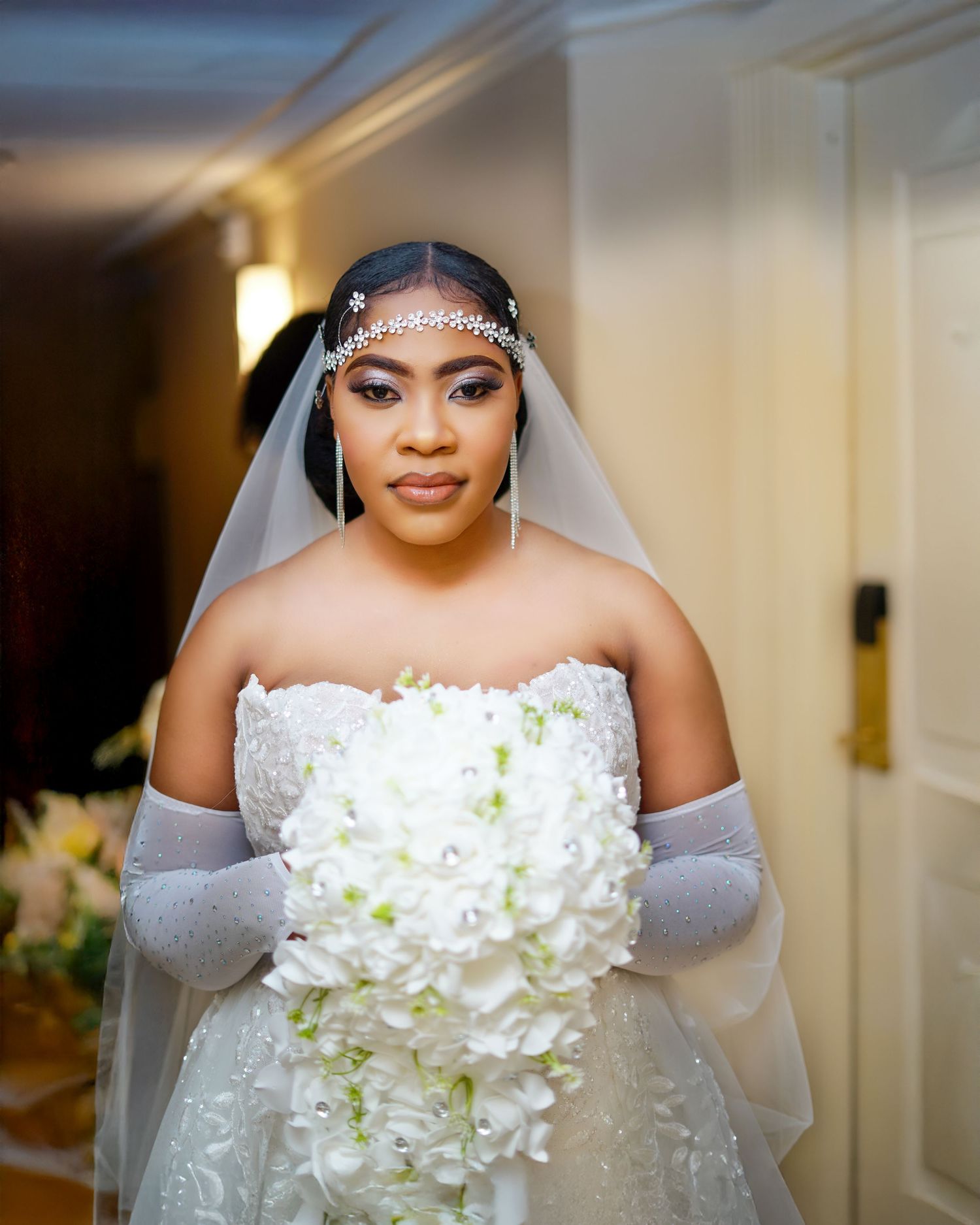 Stunning bride in strapless lace gown wearing veil and gloves, holding a cascading white floral bouquet.