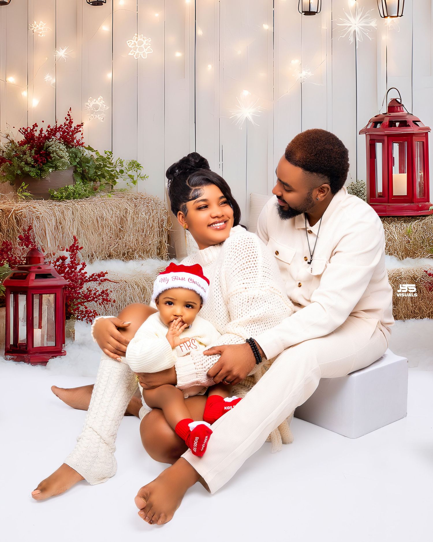 Family Christmas portrait with couple in white outfits and baby in Santa hat with festive decor.