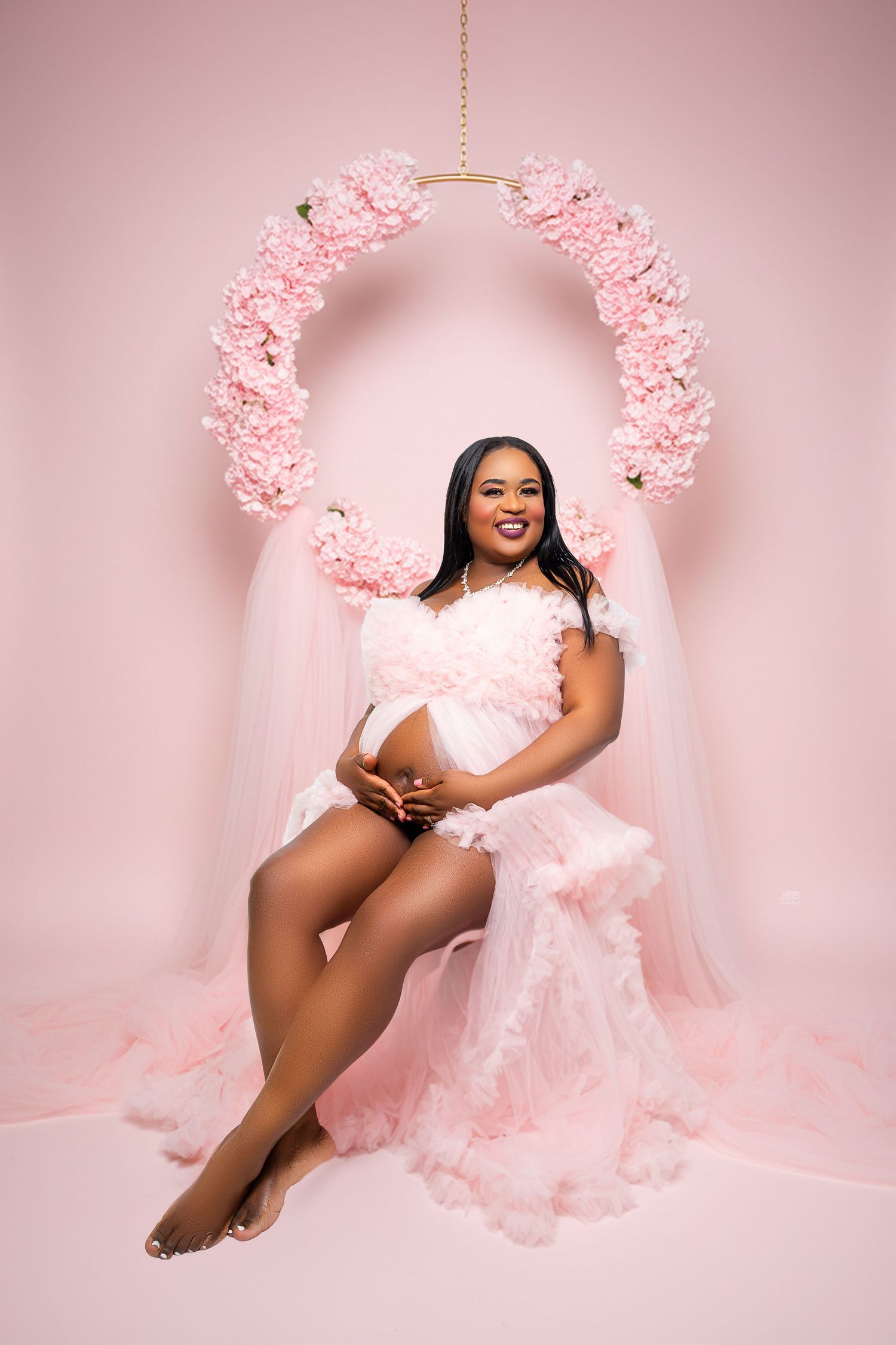 Pregnant woman in pink tulle dress seated under a floral arch in a pink studio setting.
