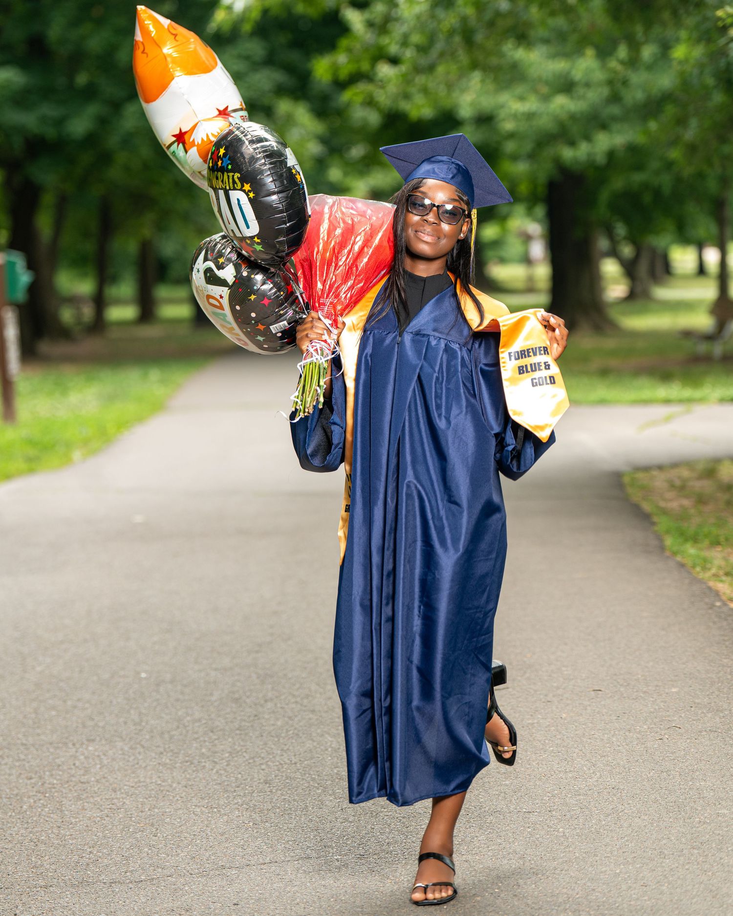 Graduate in navy blue cap and gown carrying celebration balloons outdoors.