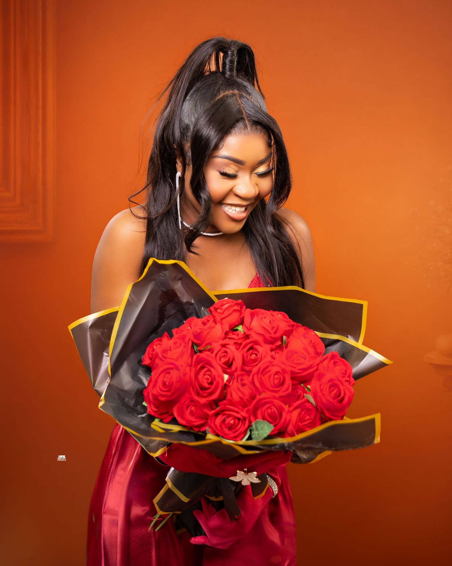 Joyful woman in red dress holds bouquet of red roses against warm orange background.