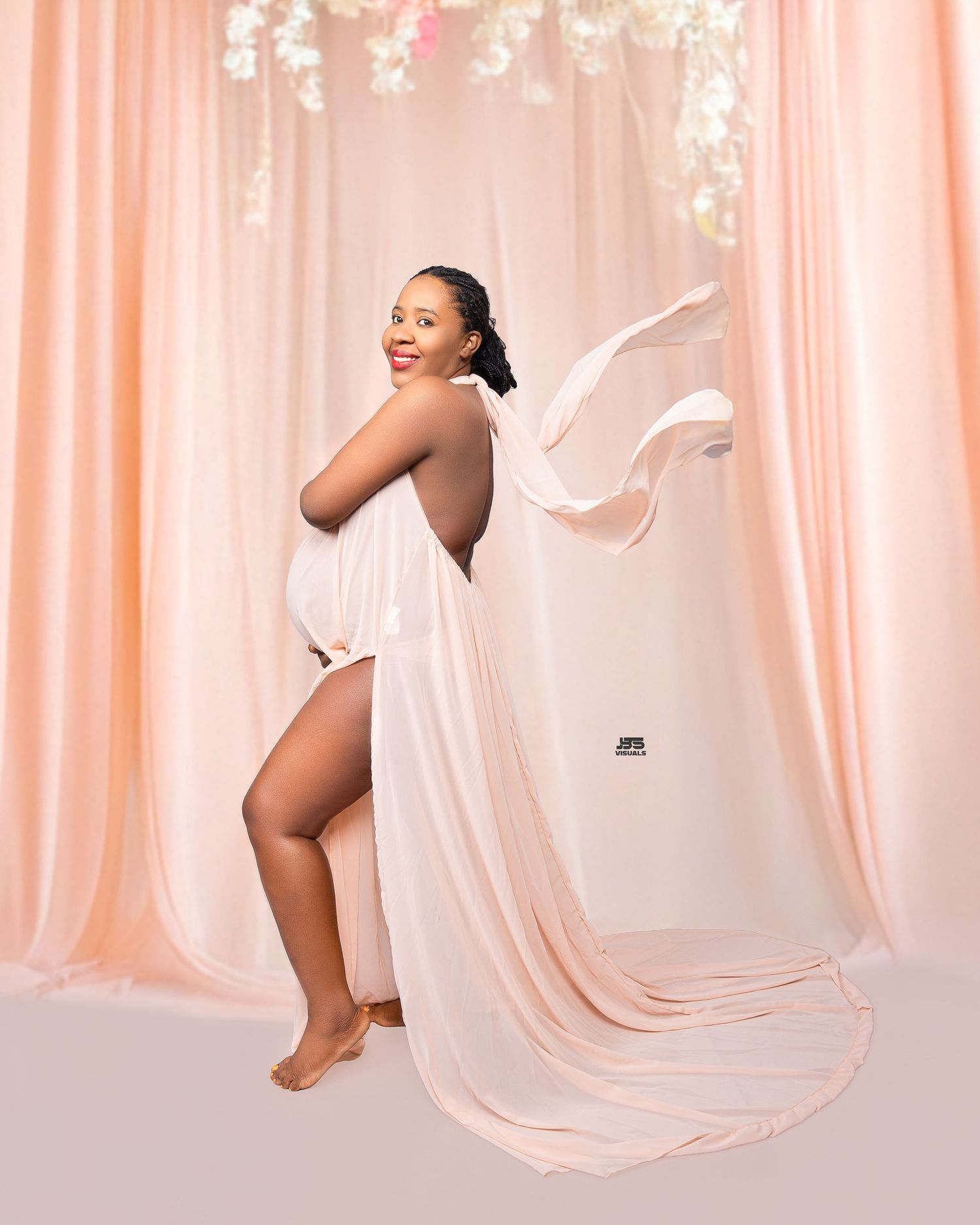 Radiant pregnant woman in flowing white sheer fabric posing against a soft pink backdrop.