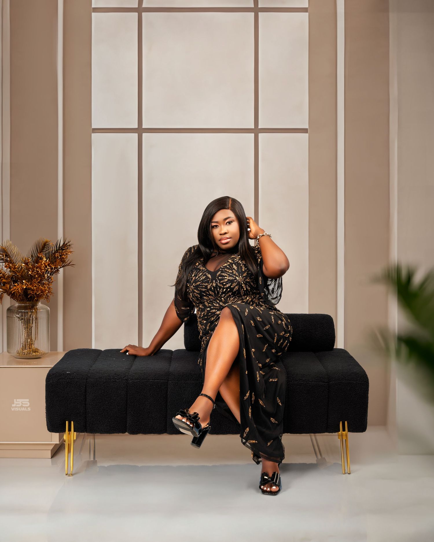 Woman in black floral dress seated on velvet bench with gold legs in elegant studio setting.