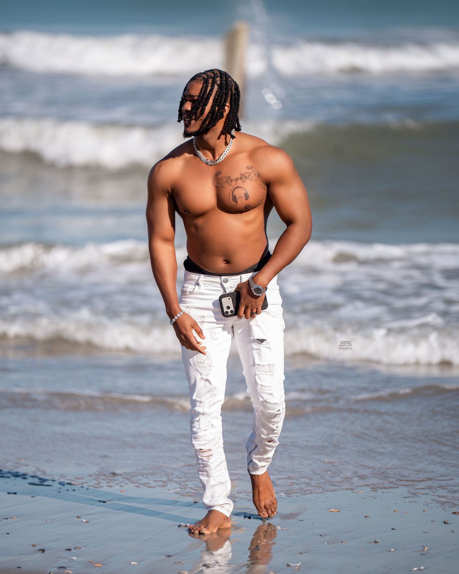 Shirtless muscular man with braids wearing white ripped jeans walking barefoot on a sunny beach with ocean waves.