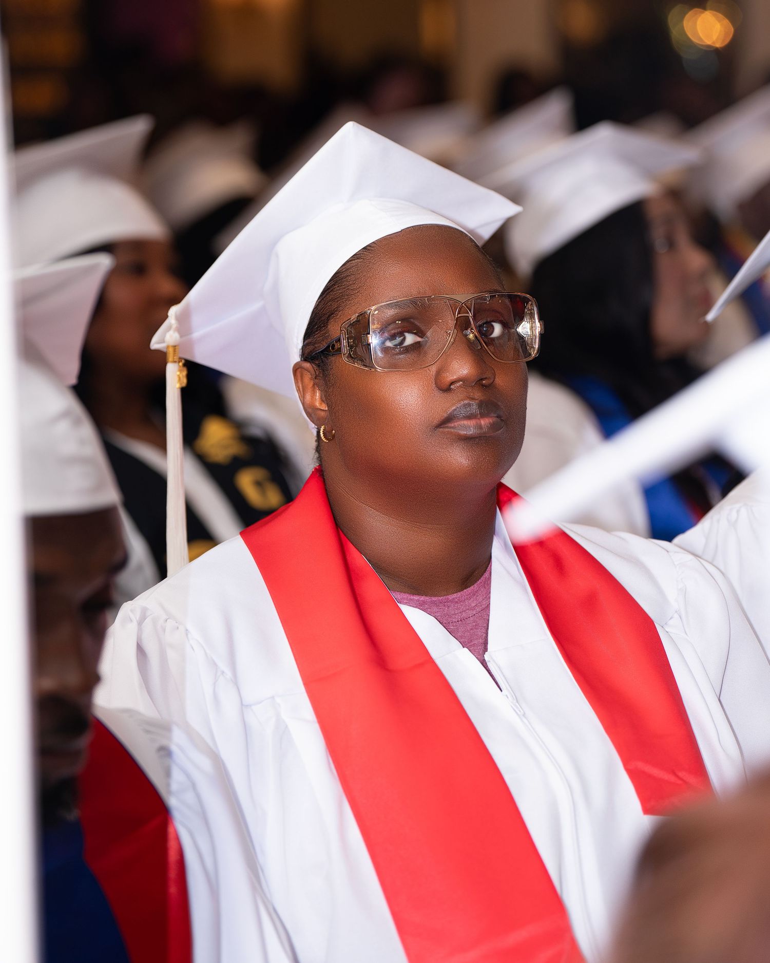Graduate in white cap and gown with red sash and clear glasses at a graduation ceremony.