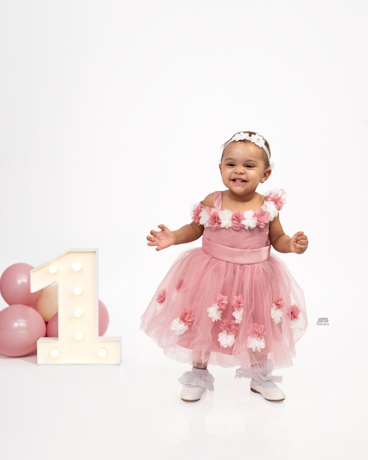 Smiling baby girl in pink floral tulle dress celebrating her first birthday with balloons and marquee number one.