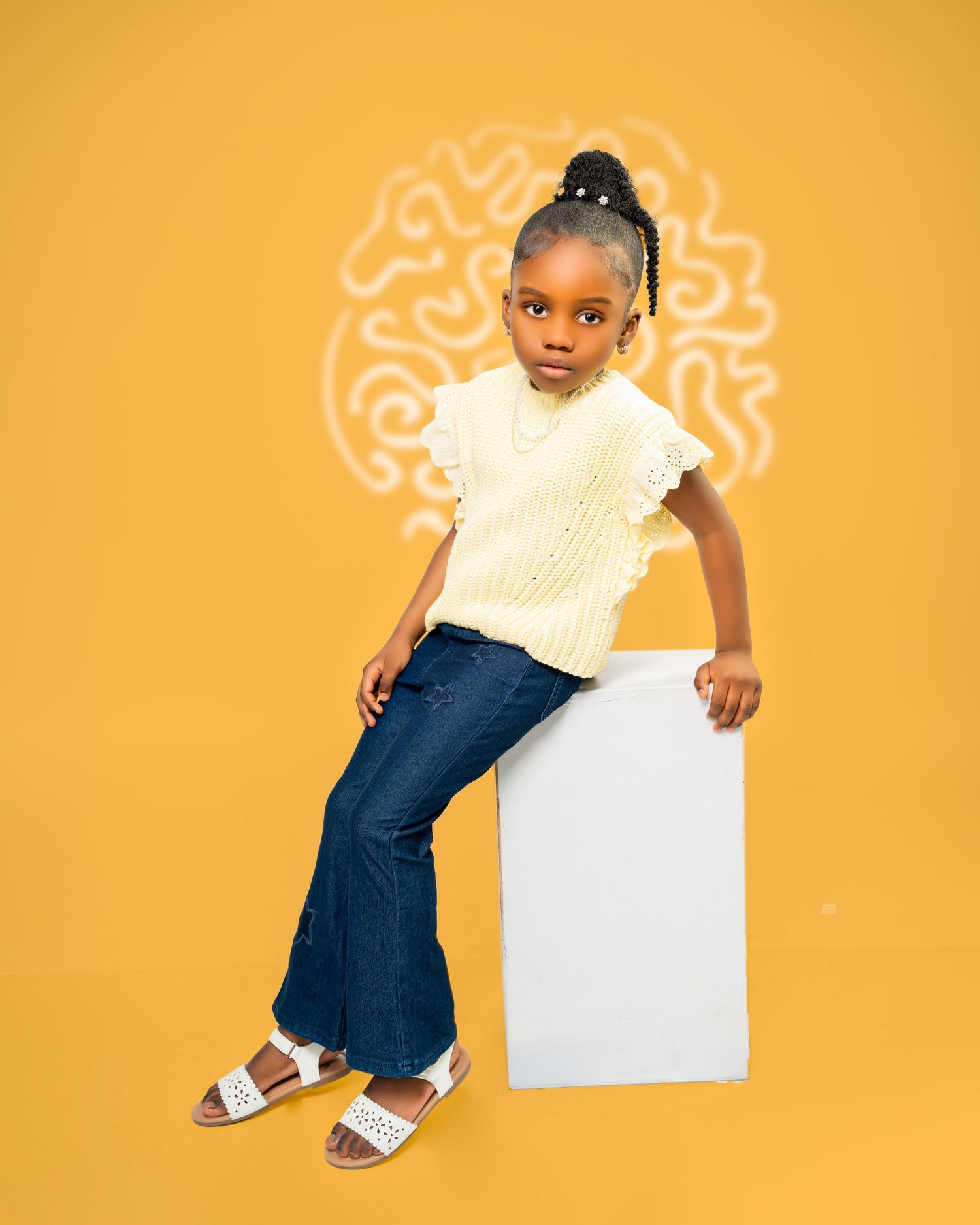 Young girl in cream knit top and jeans sitting on white cube against yellow background.