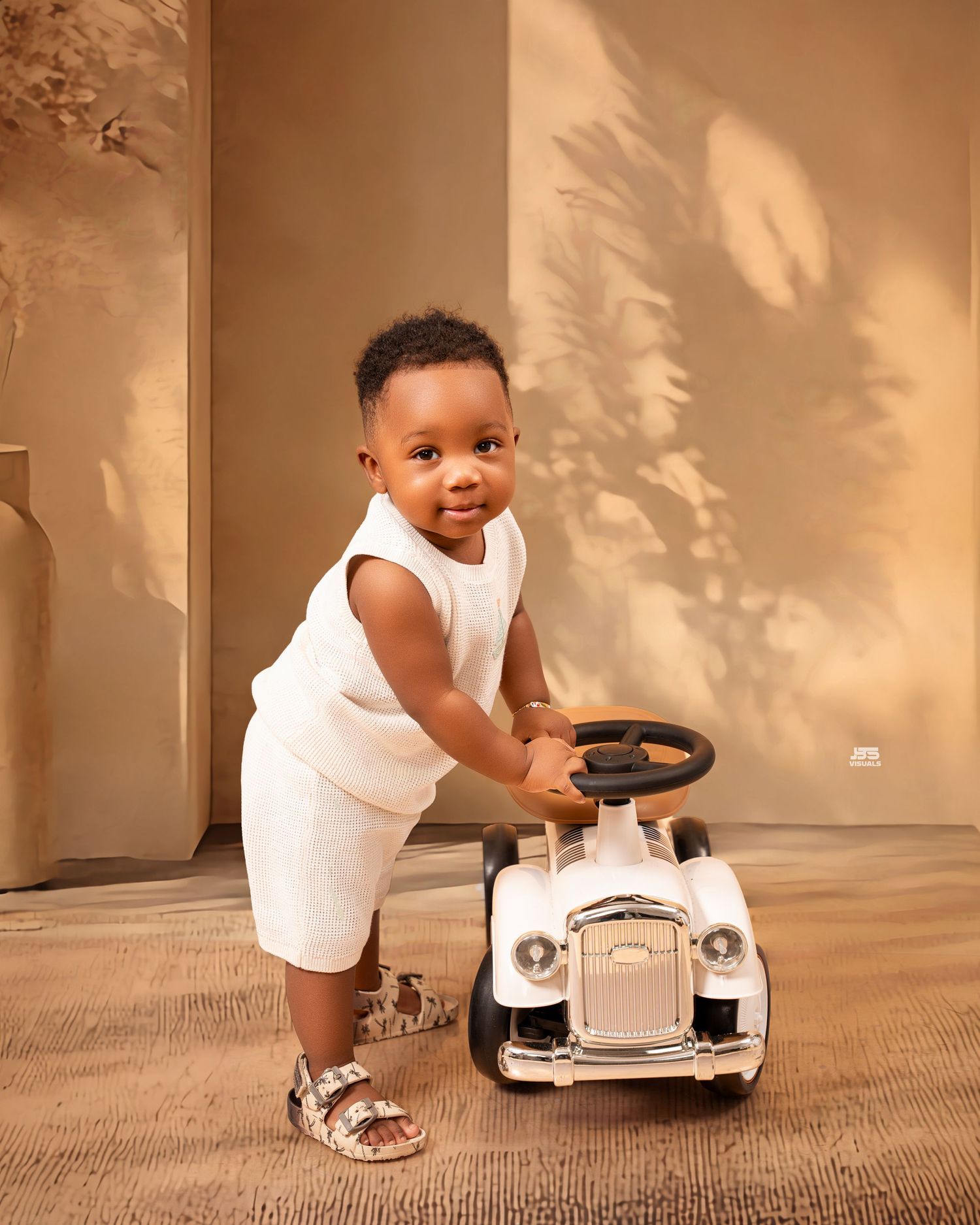Smiling toddler boy in white outfit playing with a vintage toy ride-on car on a beige floor.