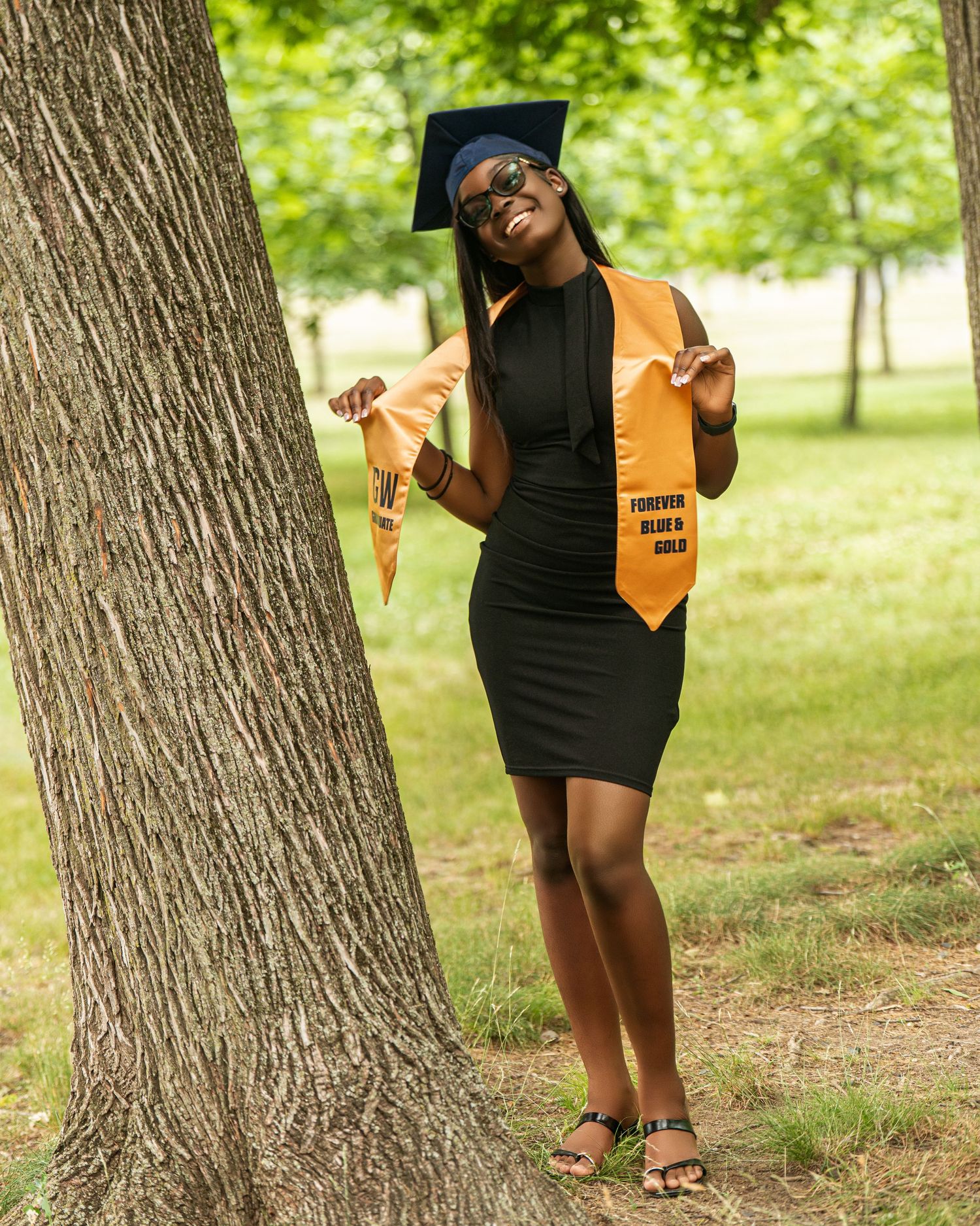 Graduate in black dress leaning against tree wearing blue cap and gold stole.