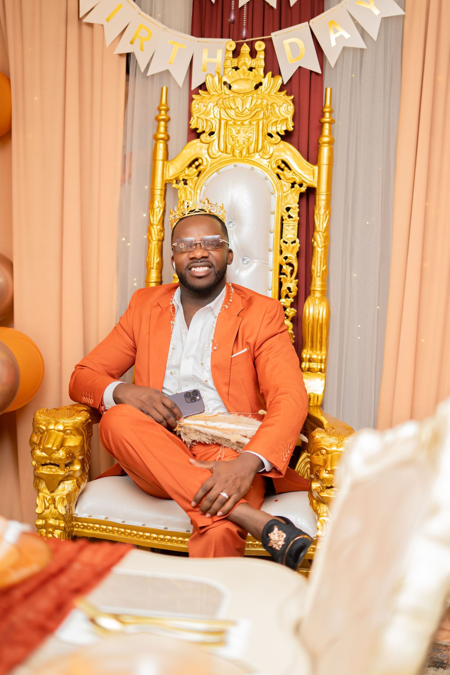 Smiling man in orange suit seated on ornate gold throne at birthday celebration.