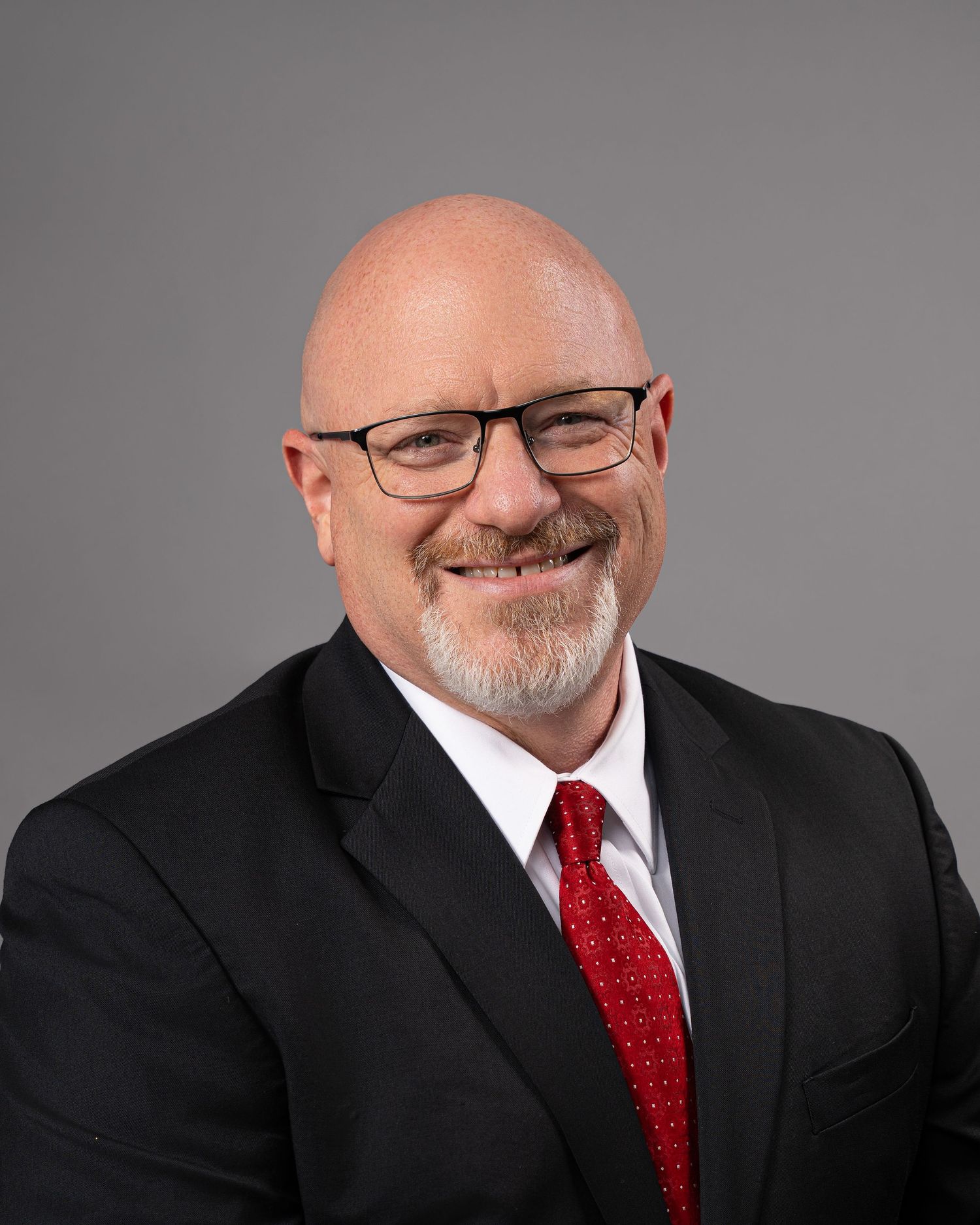 Professional headshot of a bald man with glasses, beard, wearing a black suit and red tie, smiling confidently.