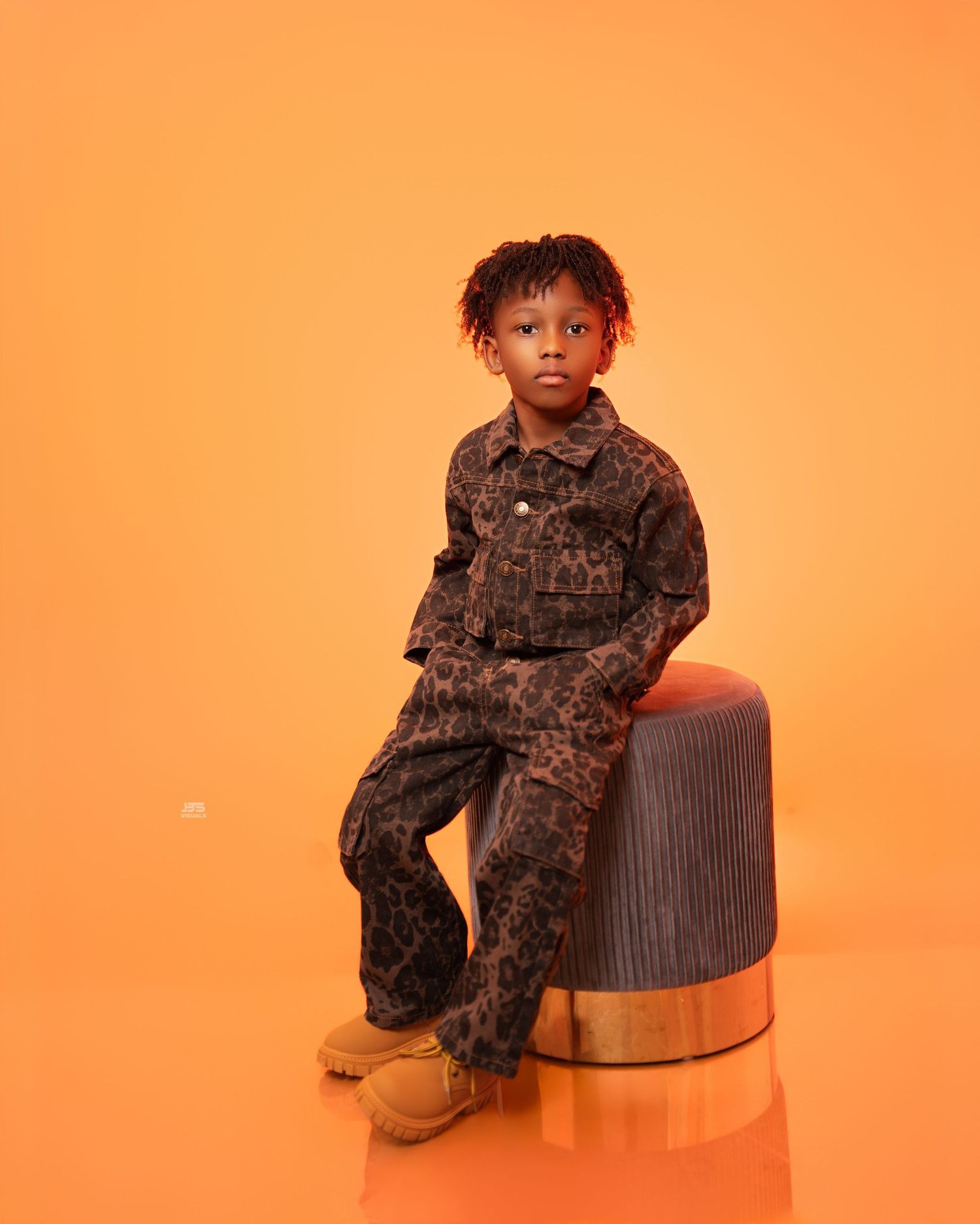Young boy in brown leopard print jumpsuit sits on a stool against an orange background in a studio photo.