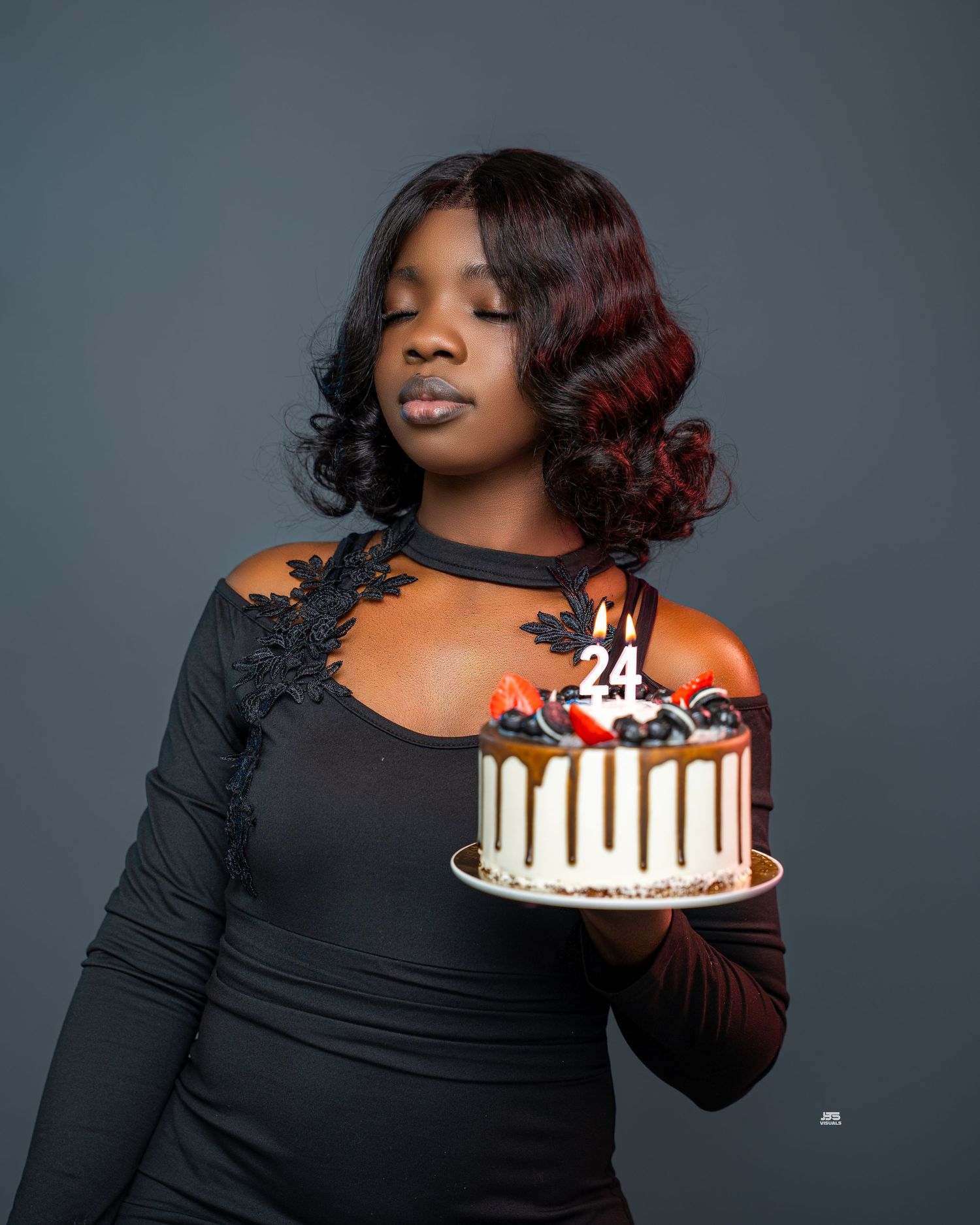 Young Black woman holding a decorated 24th birthday cake with candles in elegant black lace dress.