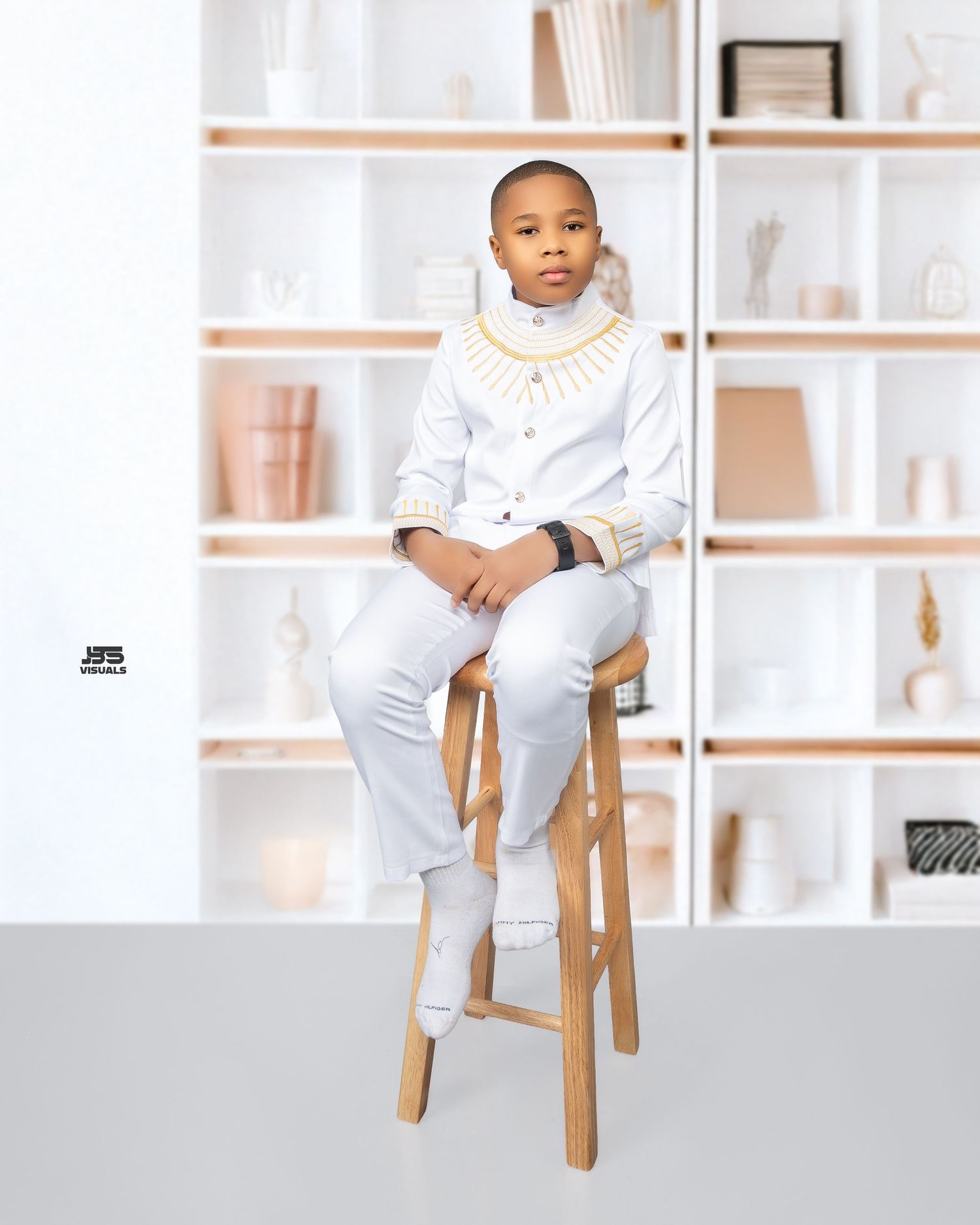 Young boy in white traditional outfit seated on wooden stool against white bookshelf backdrop.