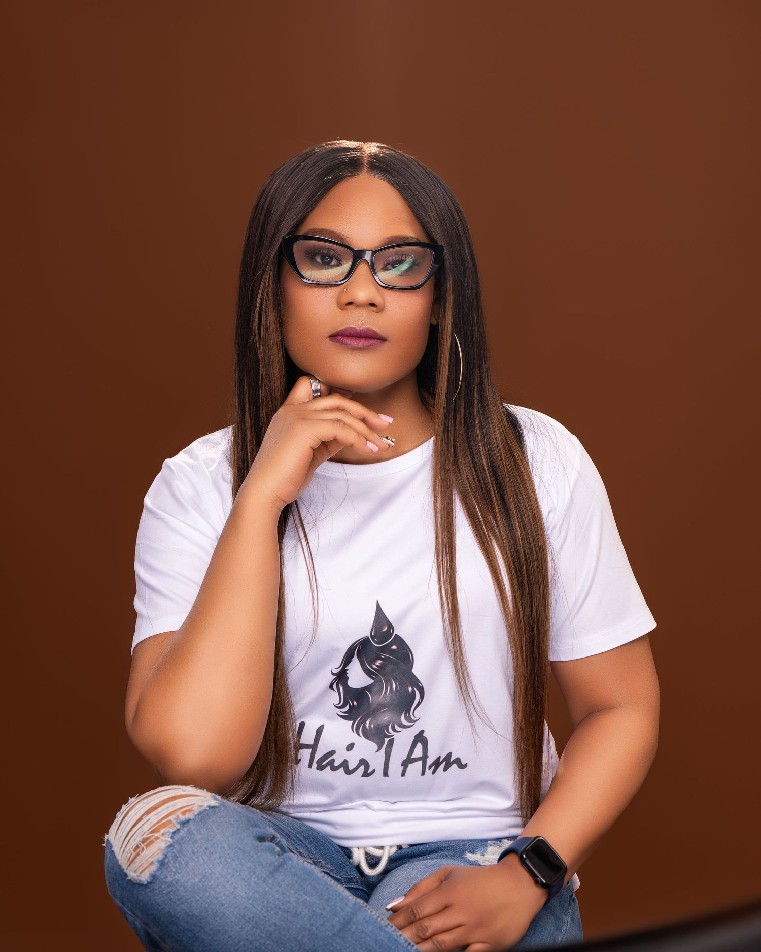 Woman in Hair I Am t-shirt and jeans sitting thoughtfully with glasses on brown background.
