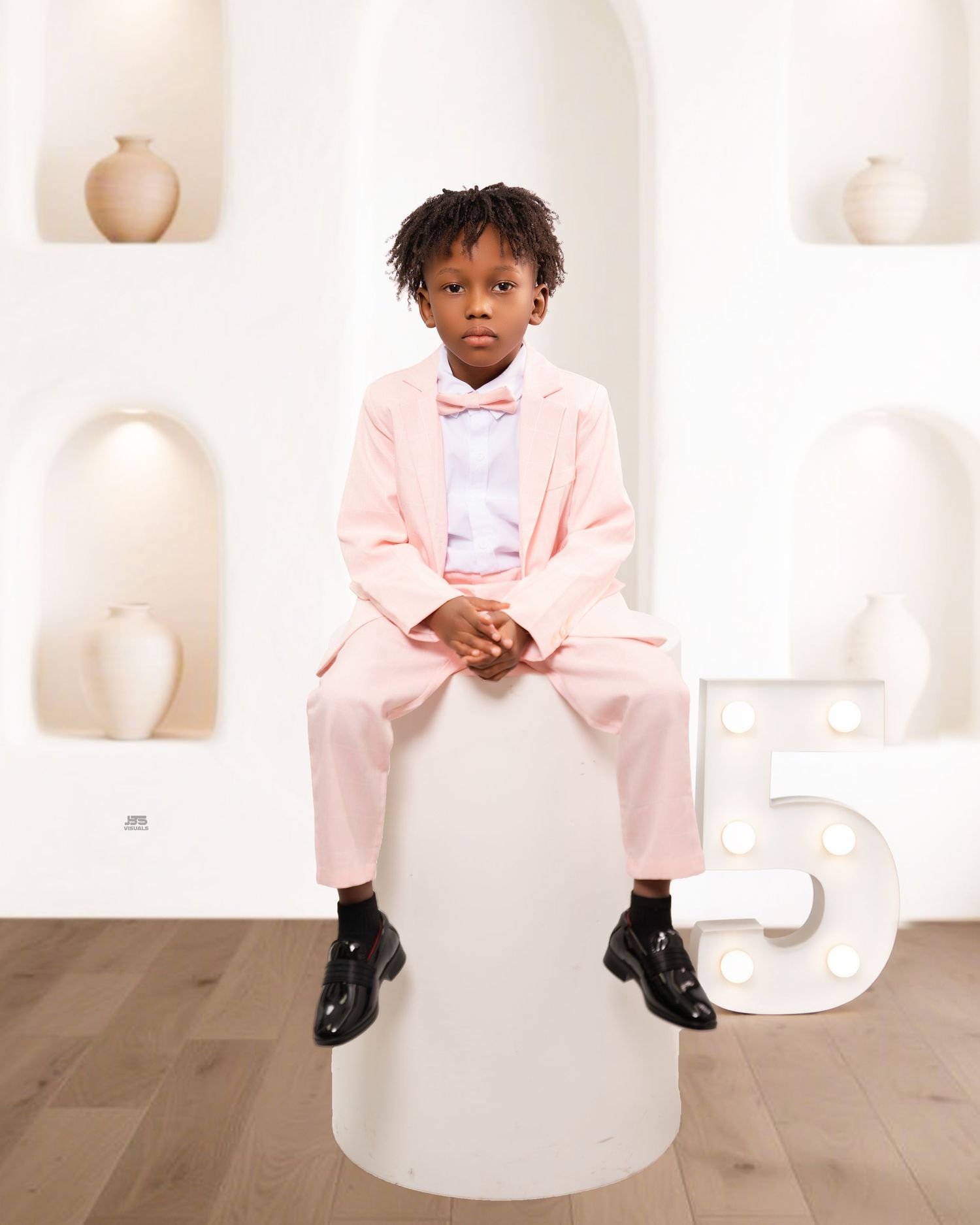 Young boy in pink suit and bow tie sits on a white pedestal beside a number 5 marquee light.