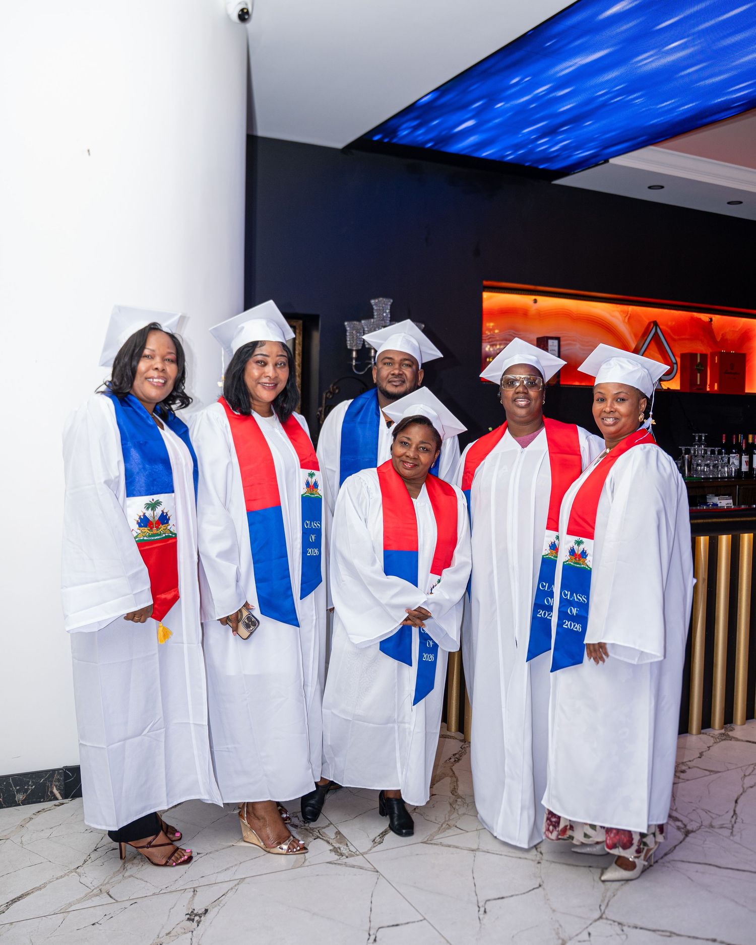 Group of graduates in white caps and gowns with Haitian flag stoles smiling together.
