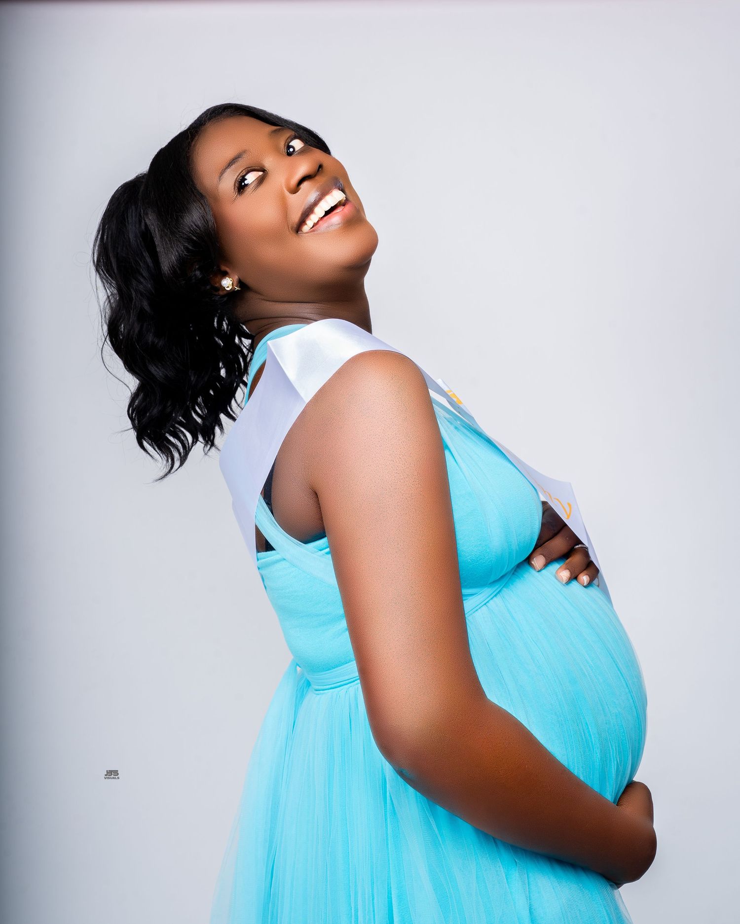 Smiling pregnant woman in aqua blue dress cradles baby bump in bright studio maternity photo.