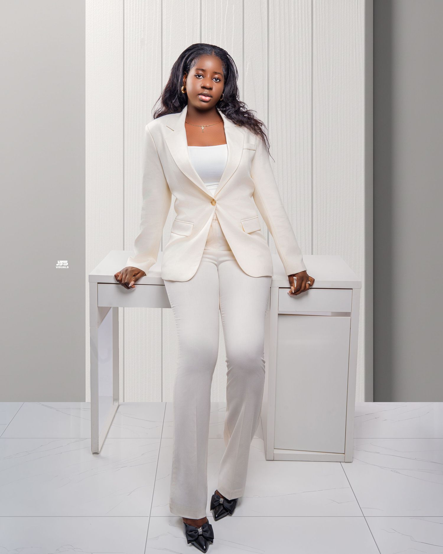 Young woman in cream white pantsuit leaning against white table in bright modern studio.