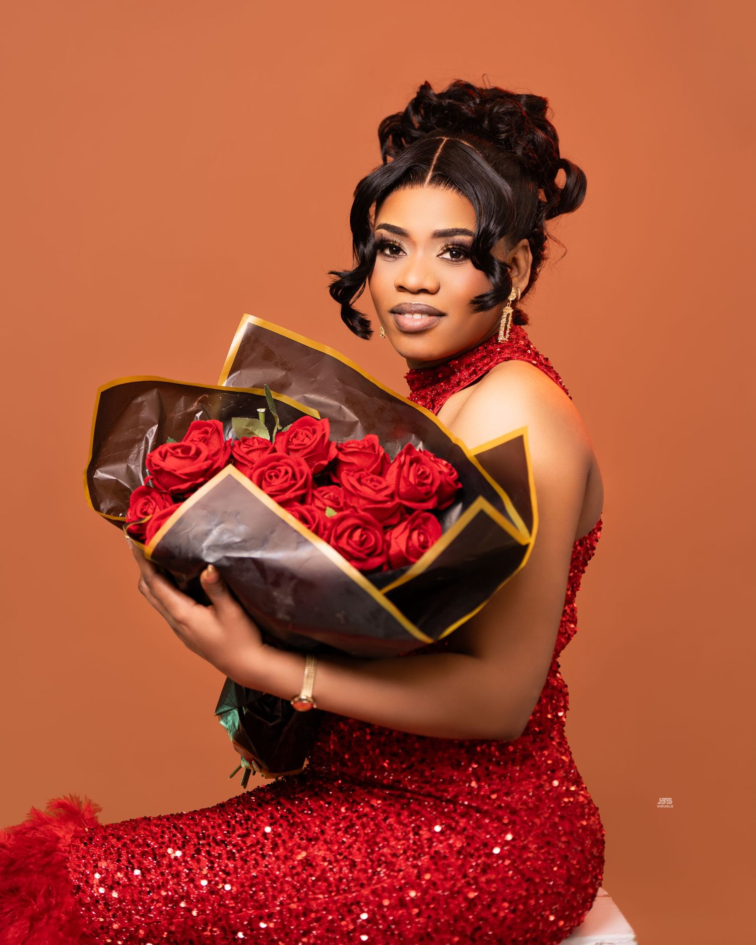 Woman in red sequined dress holds bouquet of red roses in warm orange studio photo session.