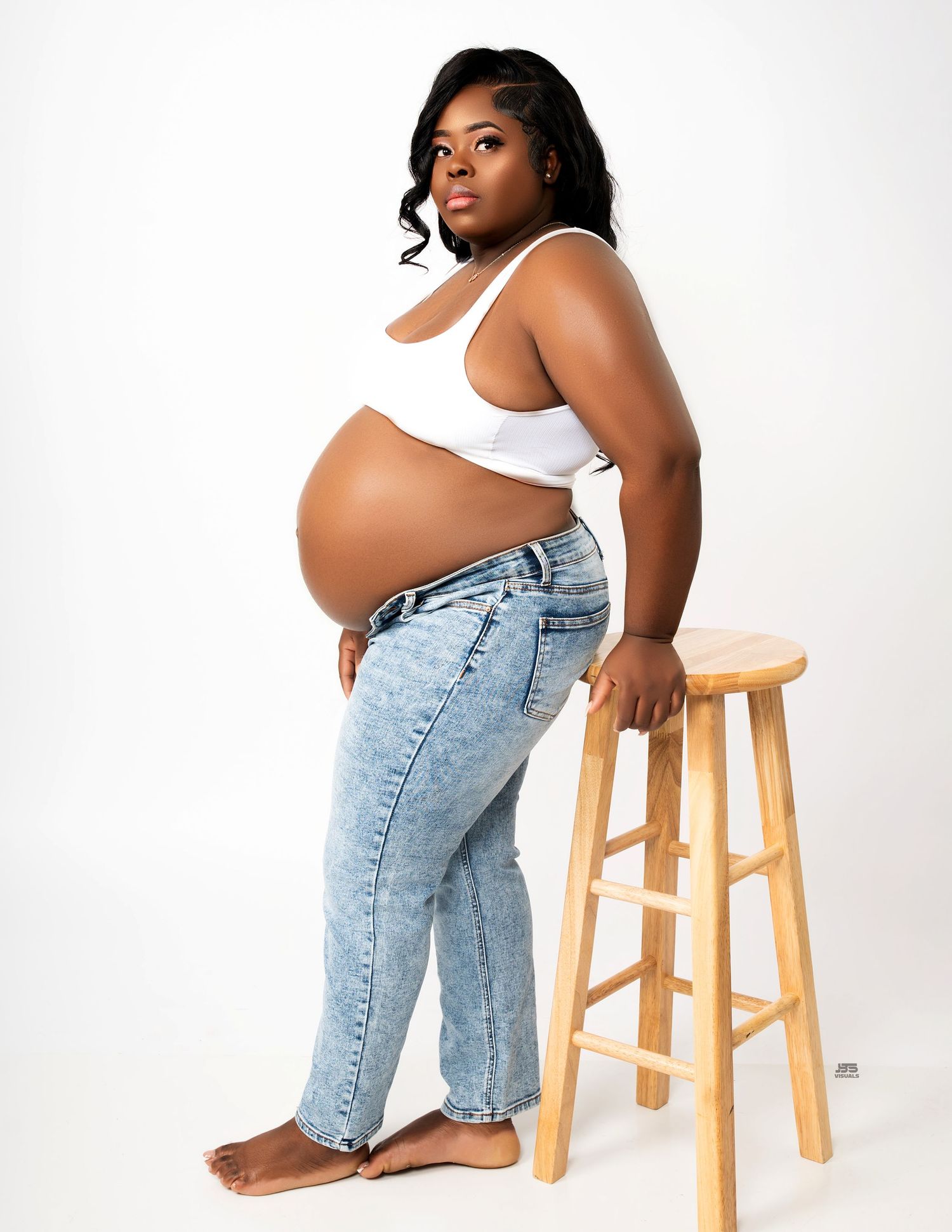Pregnant Black woman in white crop top and jeans poses beside wooden stool for maternity photo shoot.