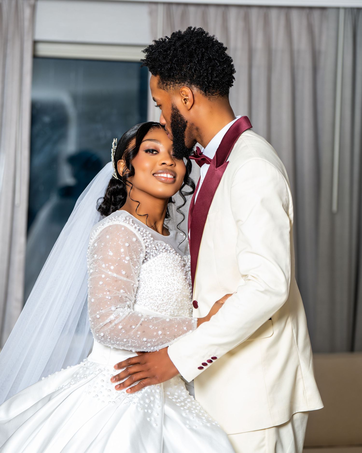 Elegant bride and groom in white and burgundy wedding attire share a romantic embrace indoors.