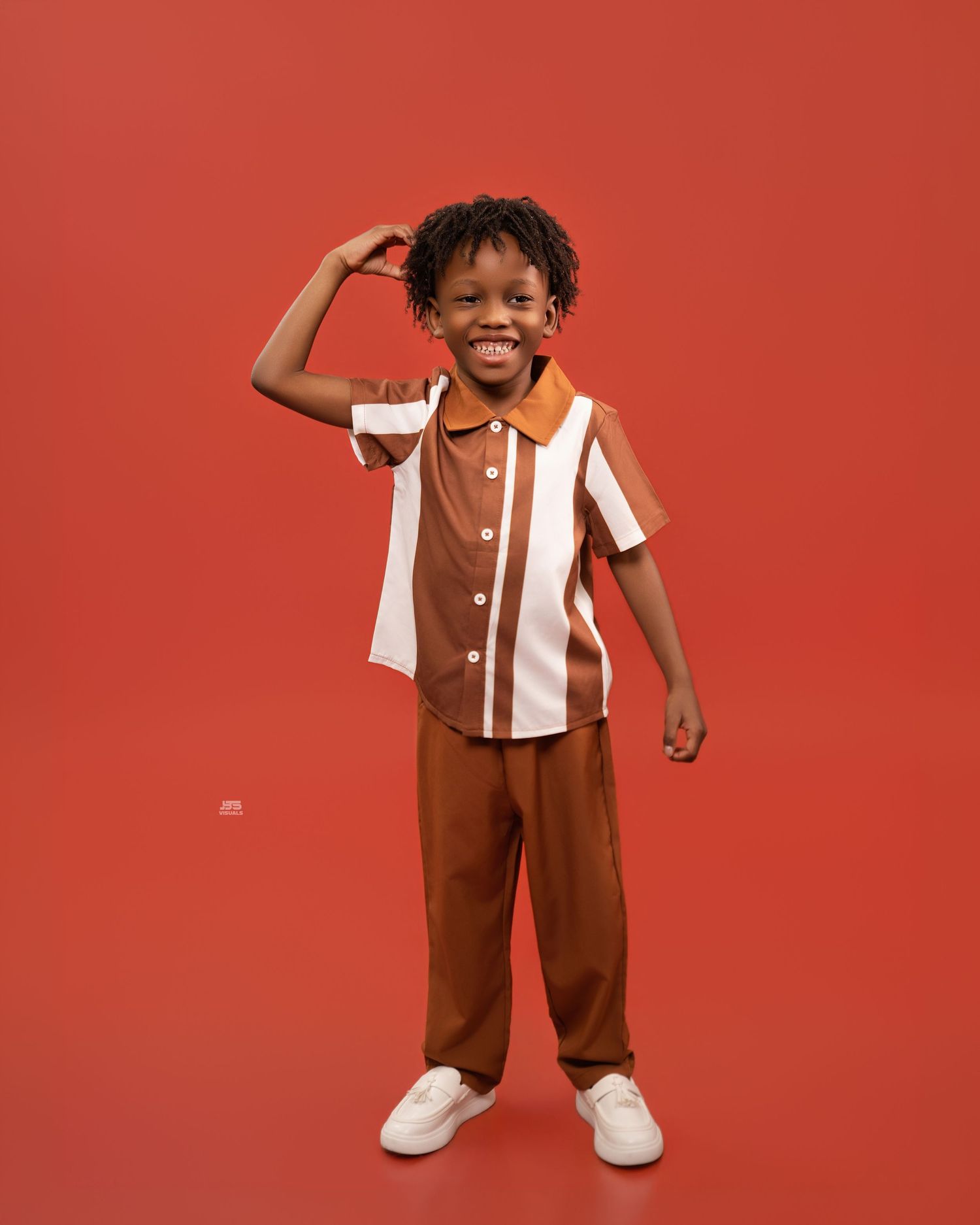 Smiling young boy in brown and white striped polo set poses confidently against a red studio background.
