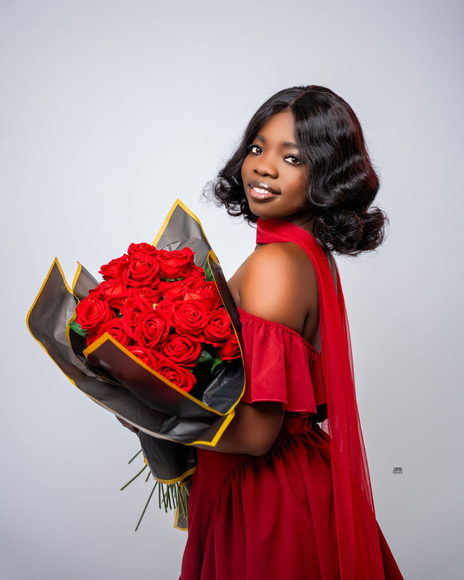 Smiling Black woman in red off-shoulder dress holding large bouquet of red roses in studio photo.