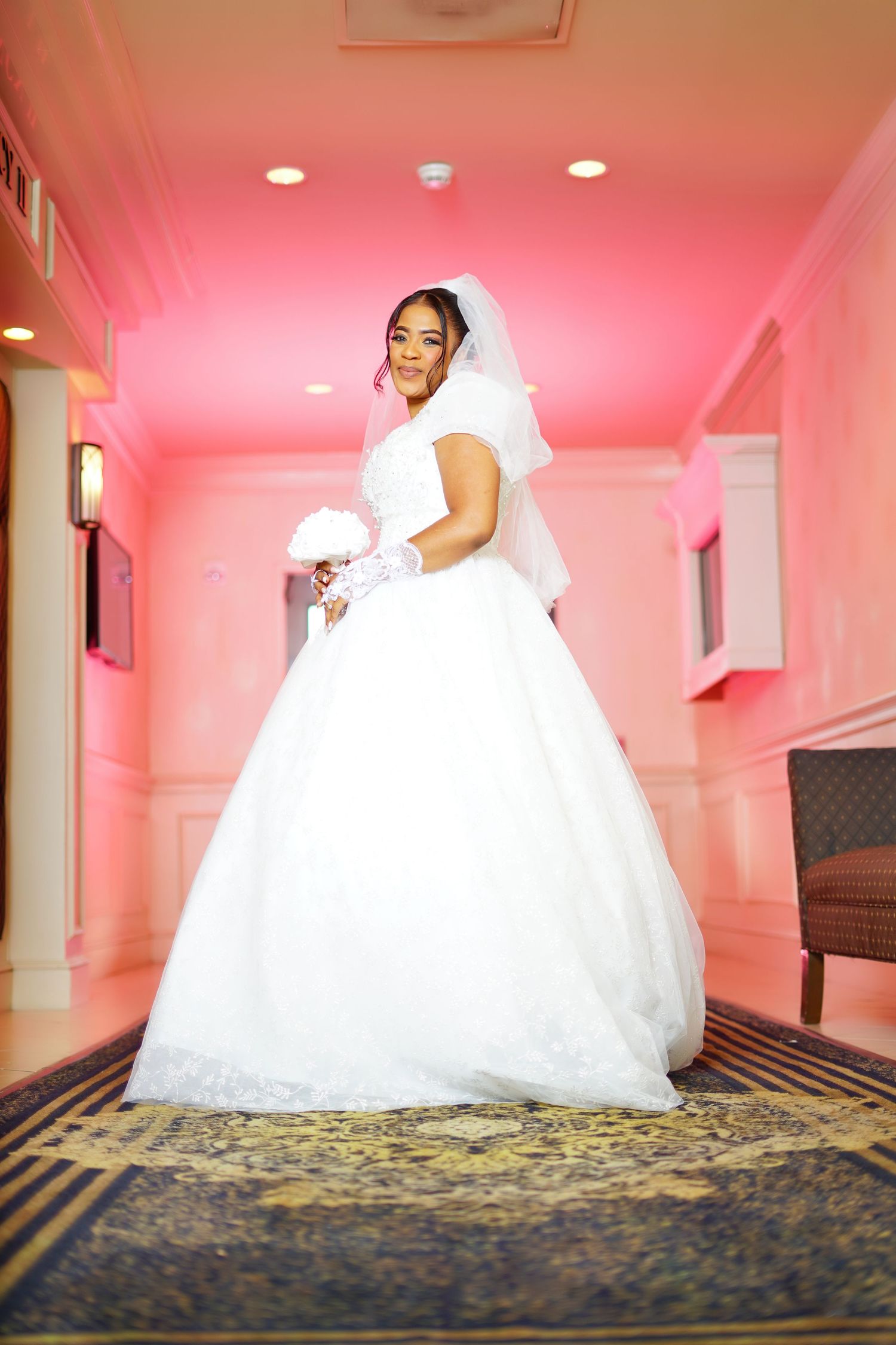 Stunning bride in full ball gown wedding dress with veil holds bouquet in pink-lit hallway.