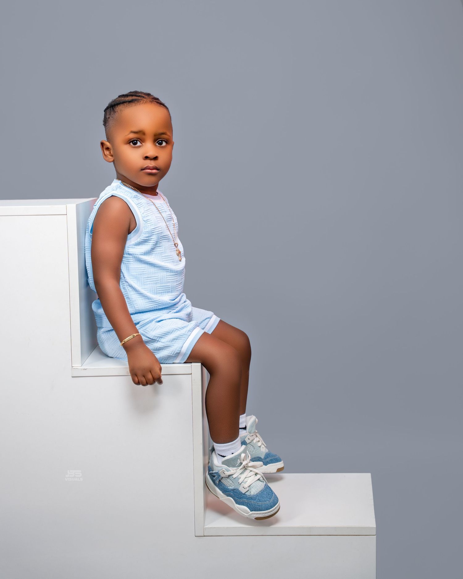Adorable young boy in light blue matching shorts set and blue sneakers sitting on white steps.