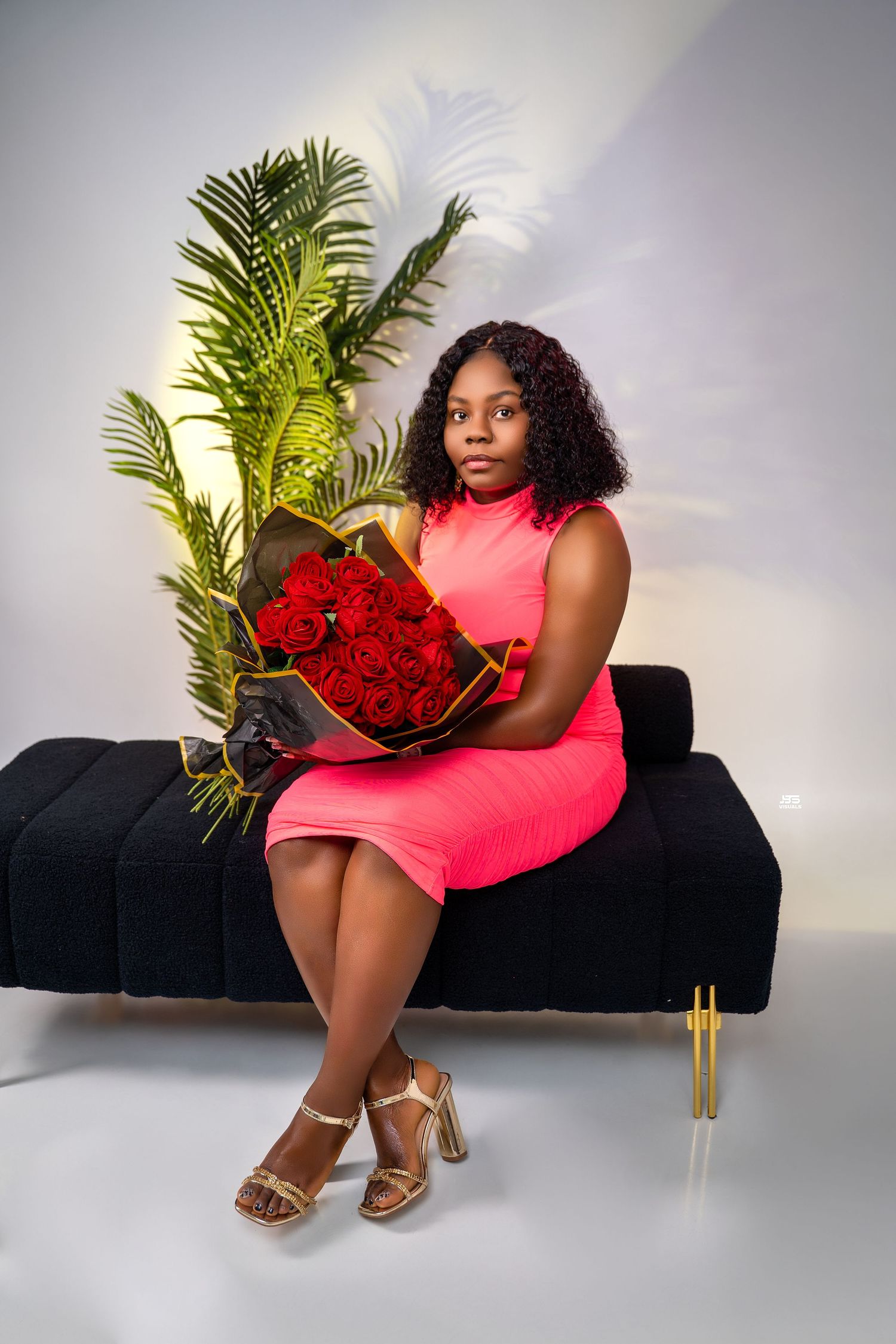Woman in pink dress holding red roses bouquet seated on black velvet bench in studio.
