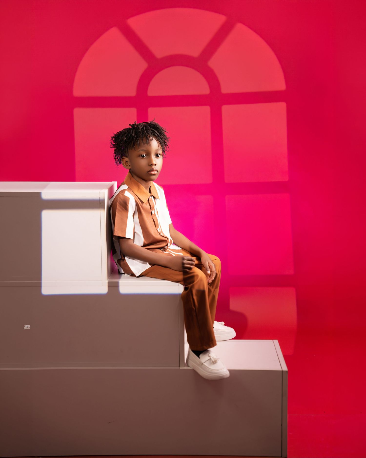 Young boy in brown and white striped outfit sits on steps against a vibrant red backdrop in studio photo.