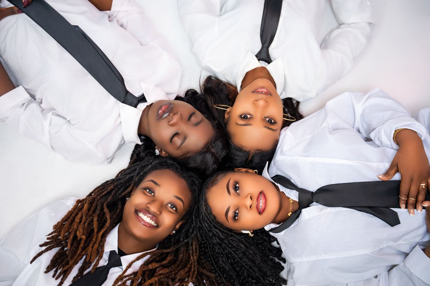 Four Black women lying in a circle wearing white shirts and black ties, shot from above.