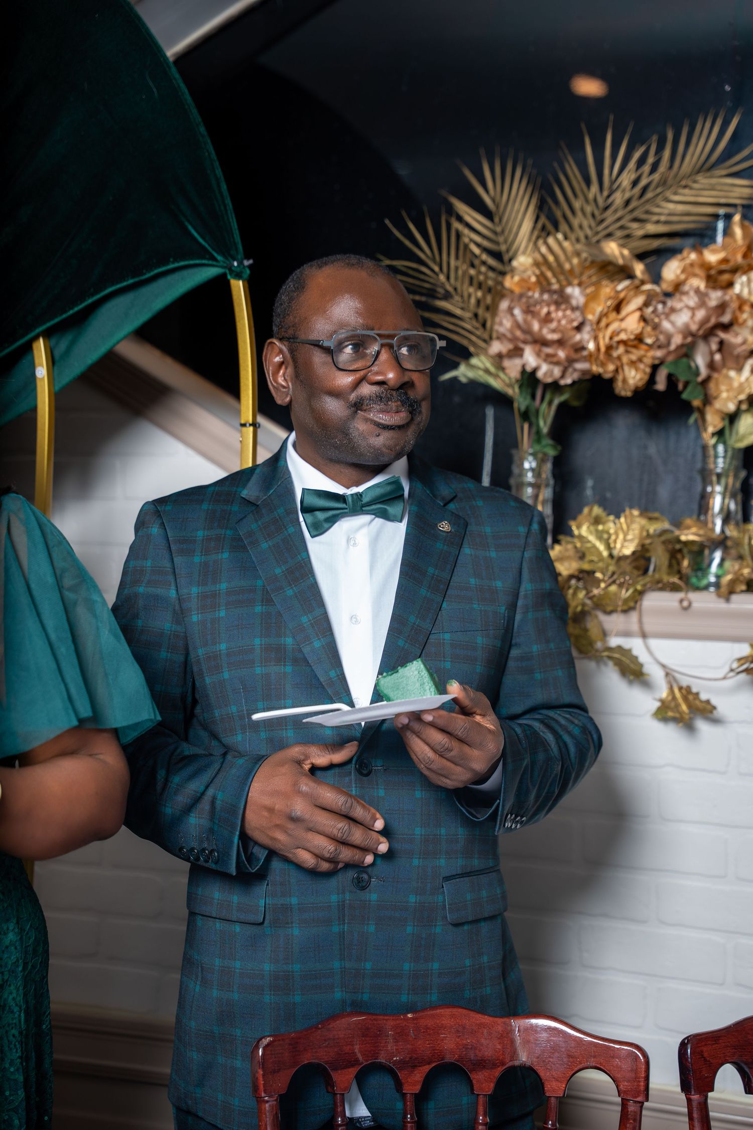 Man in green plaid suit and bow tie smiling at formal event with gold floral decorations.