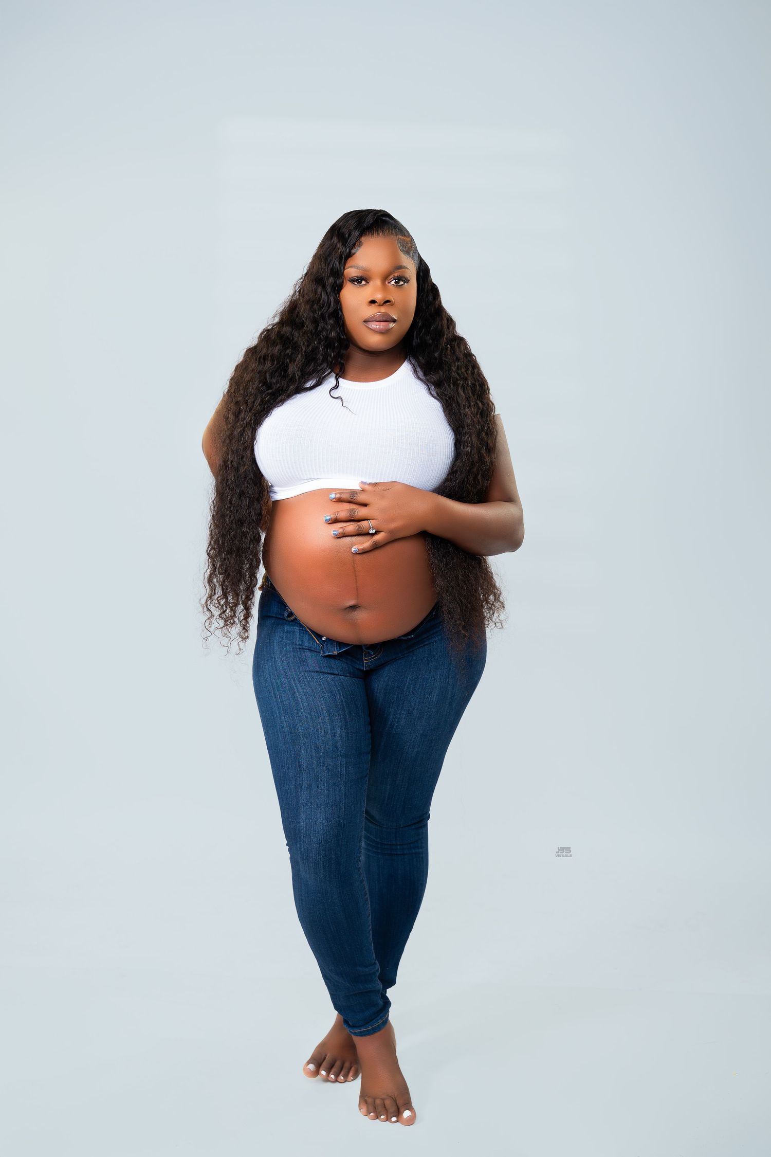 Pregnant woman in white crop top and jeans cradles bare baby bump in light blue studio setting.